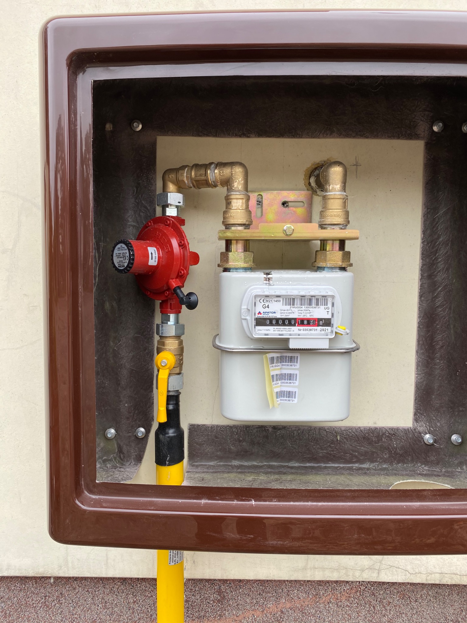 Zamknięta w brązowej obudowie instalacja gazowa z licznikiem, reduktorem ciśnienia i zaworem odcinającym, widoczne połączenia mosiężne i rury gazowe.