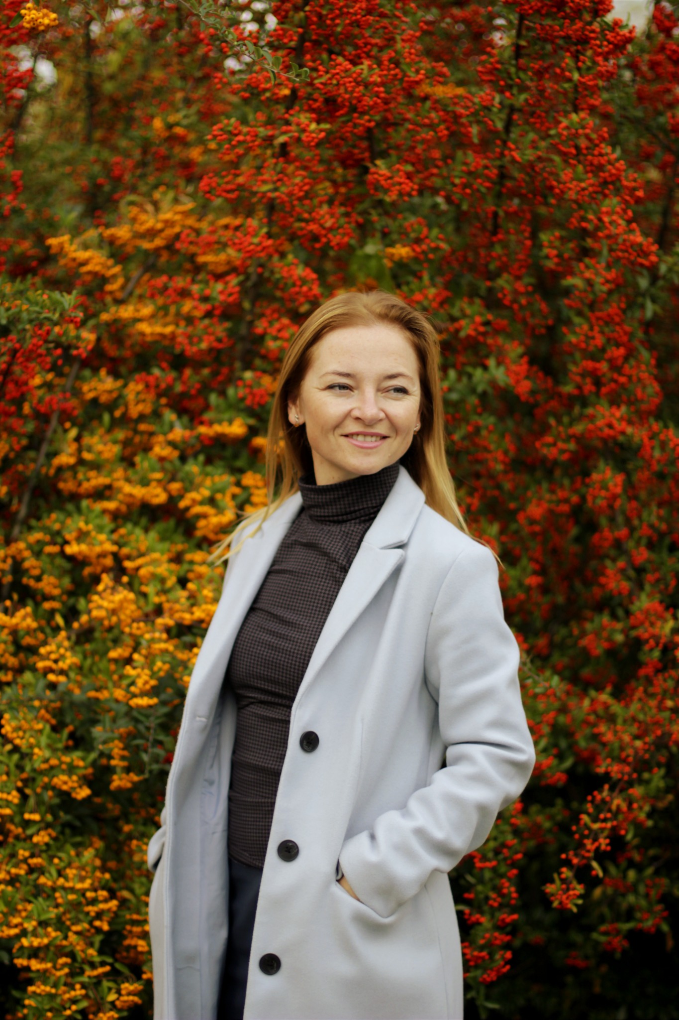 Portret kobiety w jasnym płaszczu na tle krzewu z czerwonymi i pomarańczowymi owocami, uśmiechająca się i patrząca w bok.