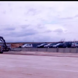 Żółta koparko-ładowarka Volvo na placu budowy, w tle zaparkowane samochody i pochmurne niebo.