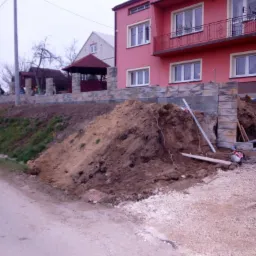 Wznoszenie muru oporowego z betonowych bloczków przy różowym, dwupiętrowym domu. W tle widoczna altana i dom. Duża pryzma ziemi przed murem, poziomica i piła spalinowa obok.