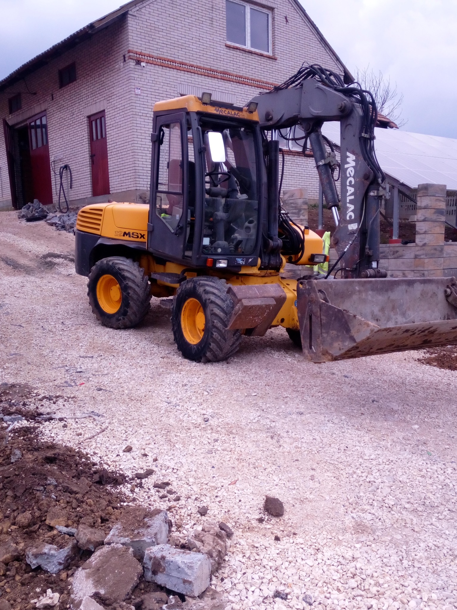 Żółto-czarna koparka Mecalac 12 MSX na żwirowym podłożu przed budynkiem z cegły, z widocznymi panelami słonecznymi w tle, sugerująca prace ziemne.