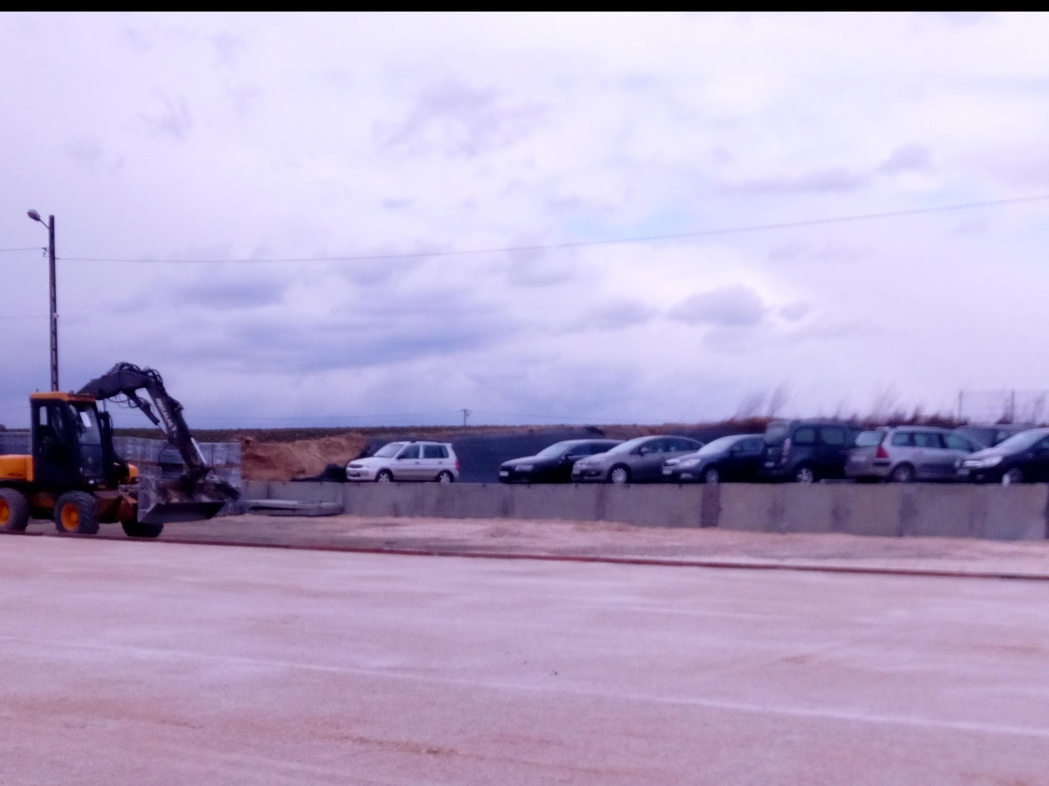 Żółta koparko-ładowarka Volvo na placu budowy, w tle zaparkowane samochody i pochmurne niebo.