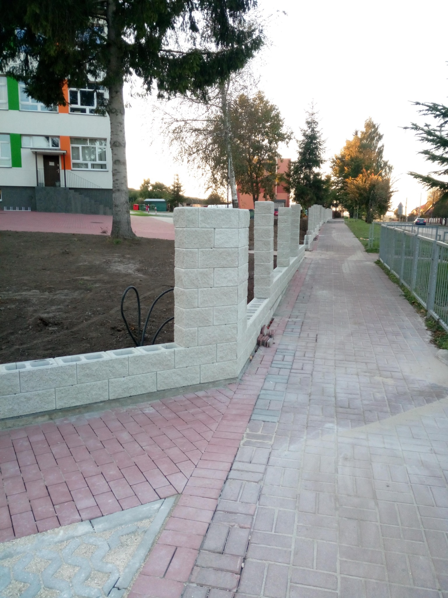 Nowo wybudowany murek z betonowych bloczków wzdłuż chodnika z kostki brukowej w odcieniach czerwieni i szarości, w tle budynek z kolorowymi akcentami i drzewa.