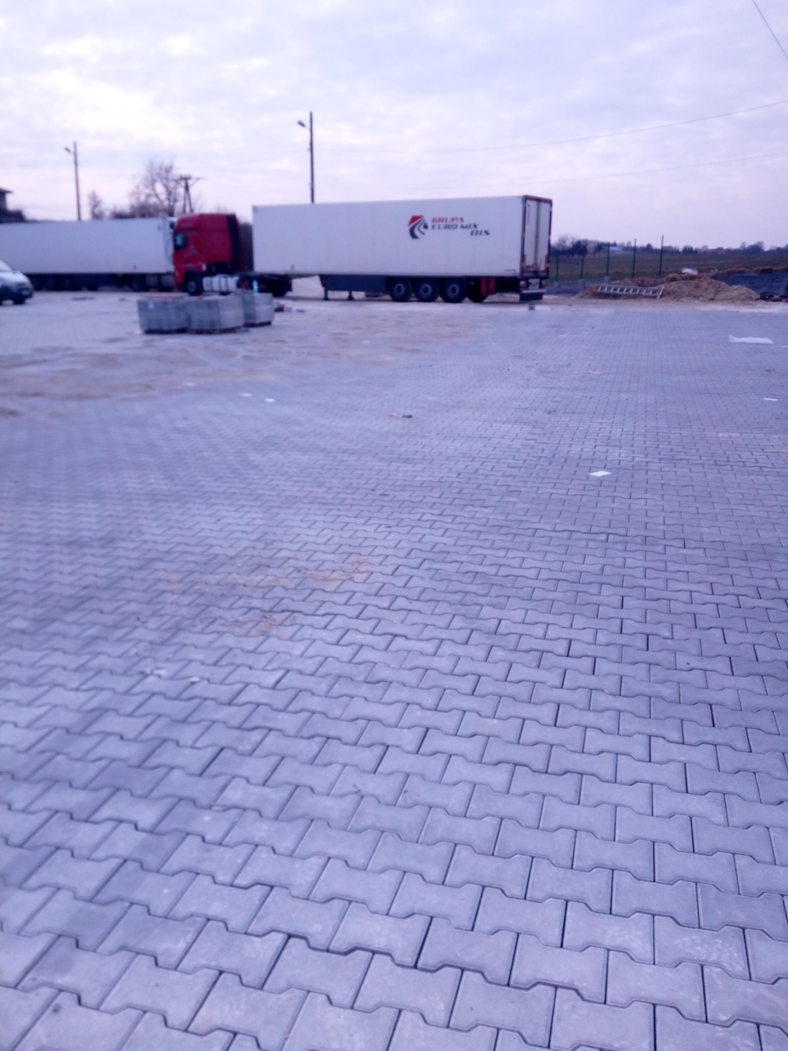 Szara kostka brukowa ułożona na placu, w tle zaparkowane ciężarówki z naczepami i sterta ziemi.