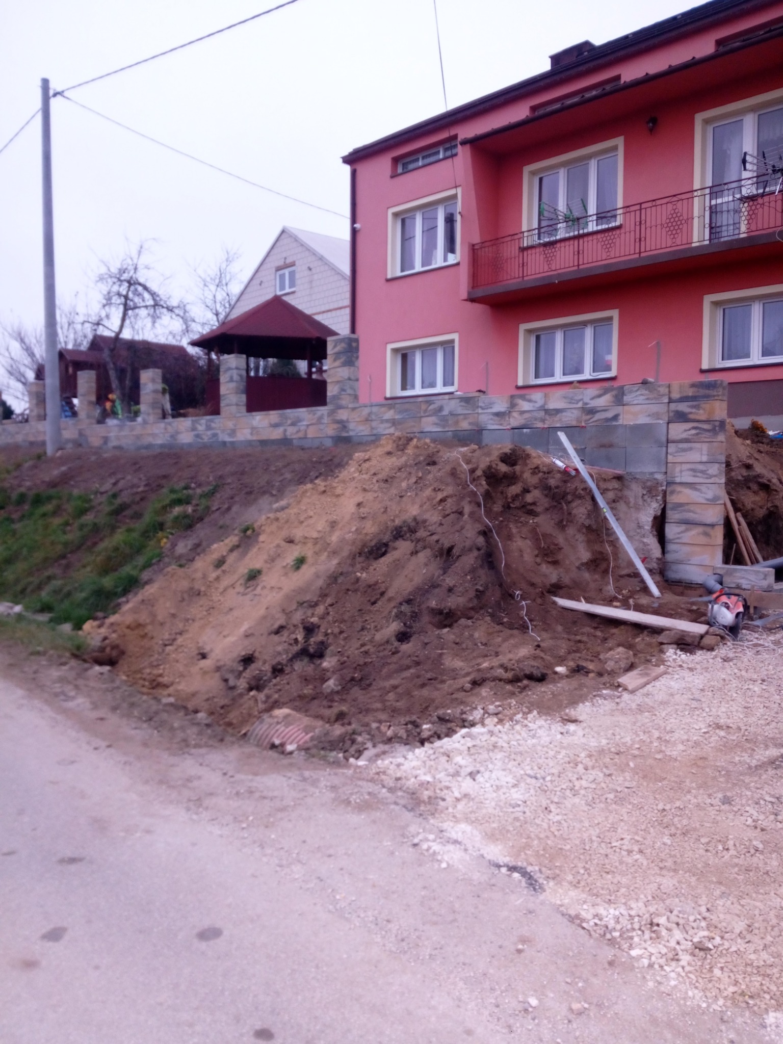 Wznoszenie muru oporowego z betonowych bloczków przy różowym, dwupiętrowym domu. W tle widoczna altana i dom. Duża pryzma ziemi przed murem, poziomica i piła spalinowa obok.