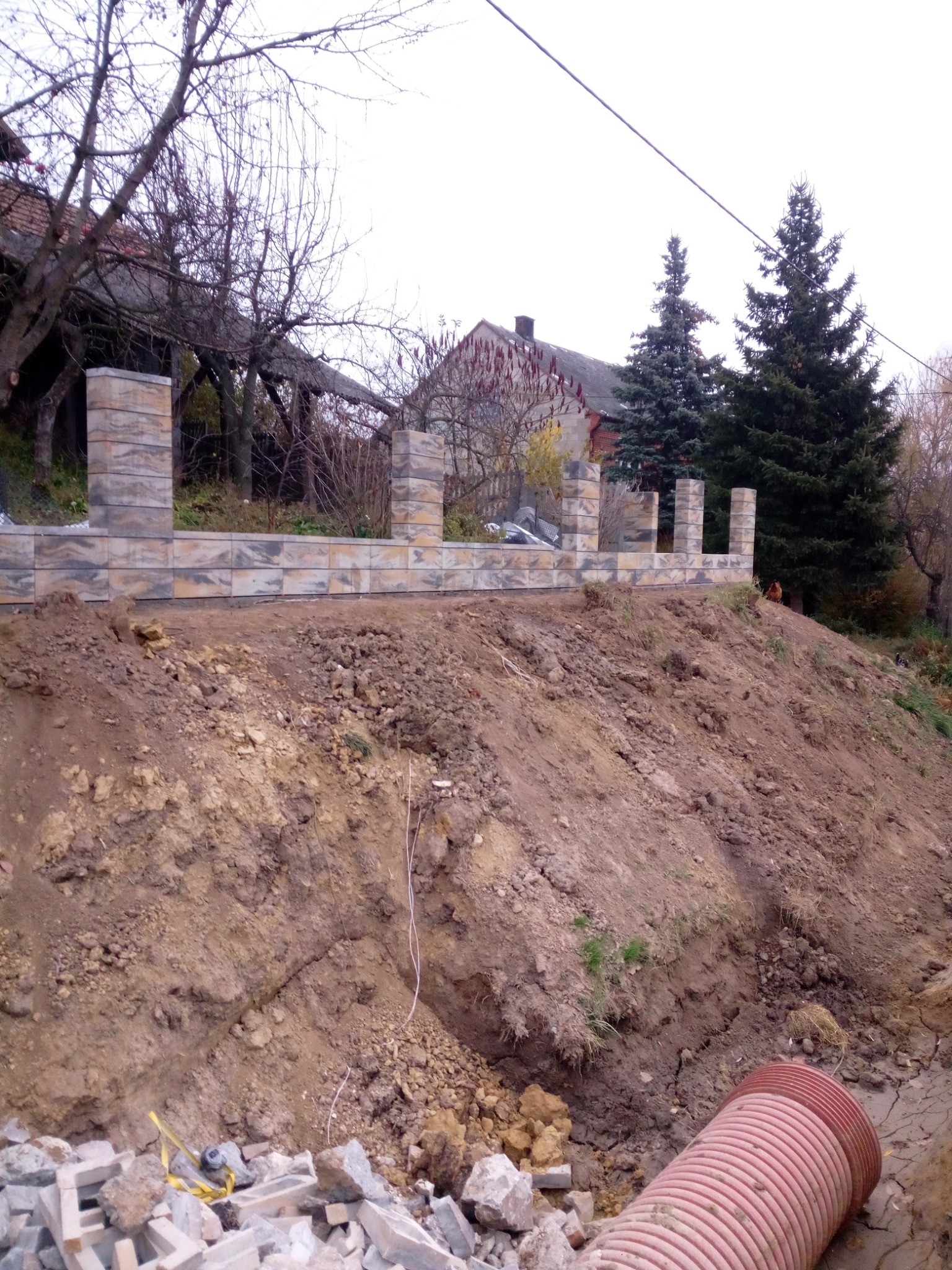 Wznoszenie ogrodzenia z prefabrykatów betonowych na nierównym terenie, widoczny rów z rurą drenarską i składowane elementy ogrodzenia, w tle domy i drzewa.