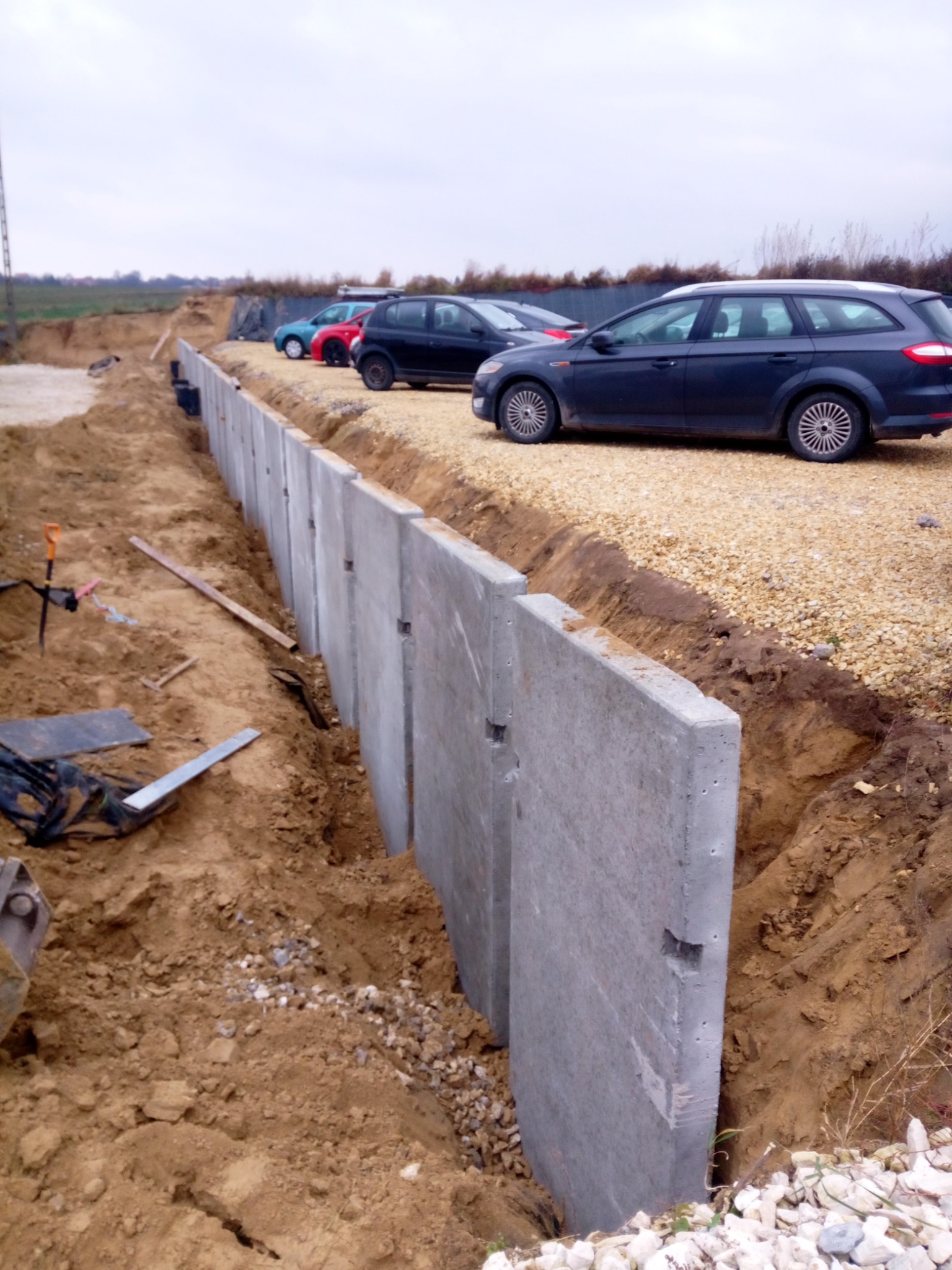 Szare betonowe panele w wykopie ziemnym, tworzące ścianę oporową; w tle zaparkowane samochody na żwirowym podłożu i pochmurne niebo.