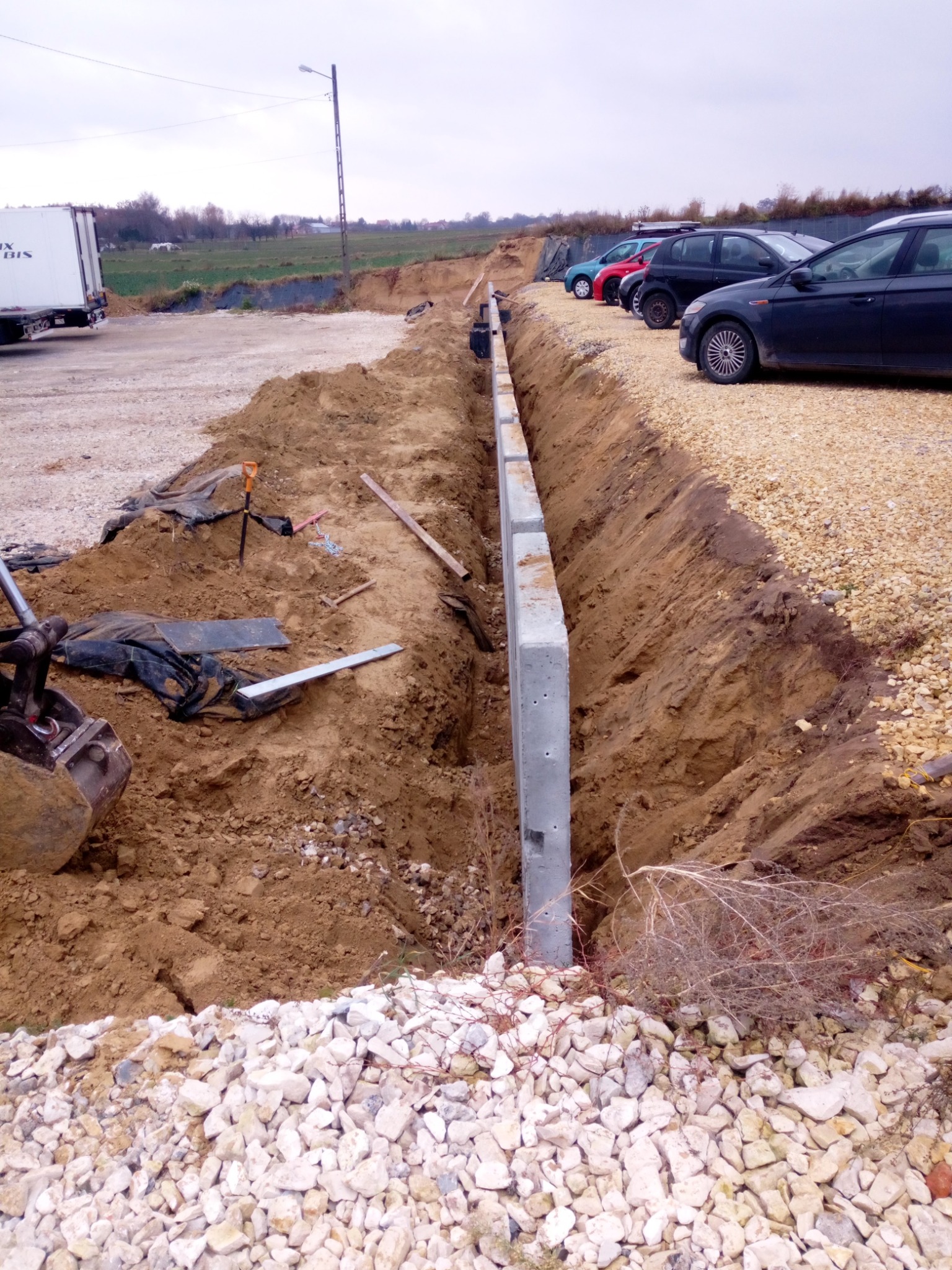 Wykop pod fundament z ustawionymi prefabrykowanymi słupami betonowymi, widoczne narzędzia i maszyna budowlana w tle, parking z samochodami.