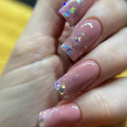 Detaliczne ujęcie dłoni z kwadratowymi paznokciami w odcieniach pudrowego różu, zdobionymi brokatem holograficznym i delikatnymi refleksami światła.