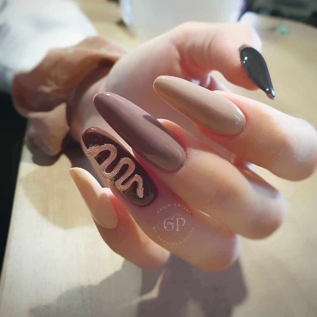 Dłoń z długimi, migdałowymi paznokciami w odcieniach beżu i brązu, z jednym paznokciem ozdobionym złotym motywem węża i logo artystki.