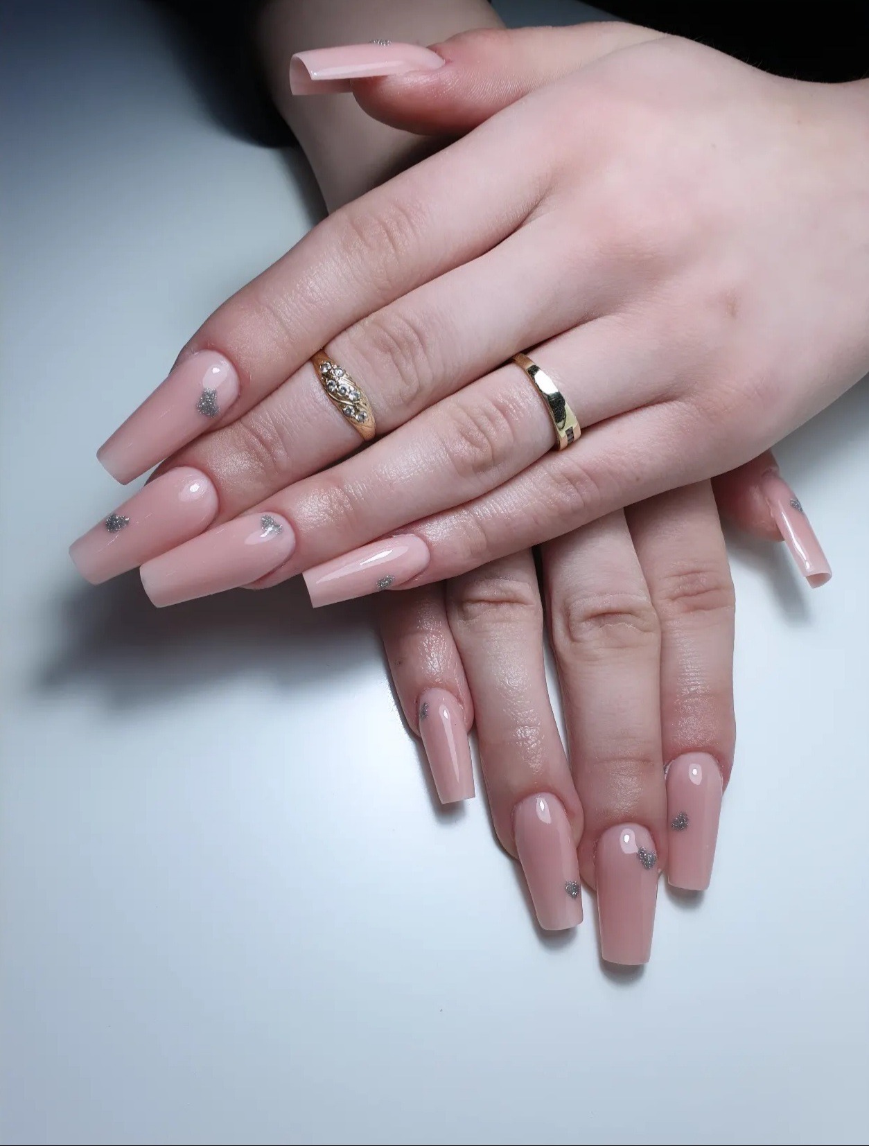 Dłonie z długimi, kwadratowymi paznokciami w odcieniu pudrowego różu, ozdobionymi srebrnymi drobinkami w kształcie serc, na palcach widoczne złote pierścionki.
