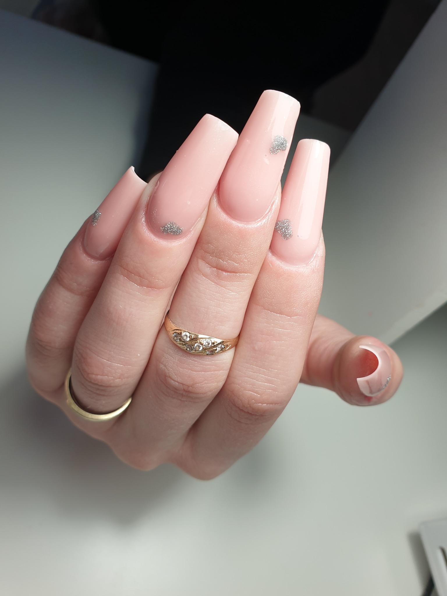 Dłoń z długimi, kwadratowymi paznokciami w odcieniu pudrowego różu, ozdobionymi srebrnym brokatem w kształcie chmurek, z pierścionkami na palcach na białym tle.