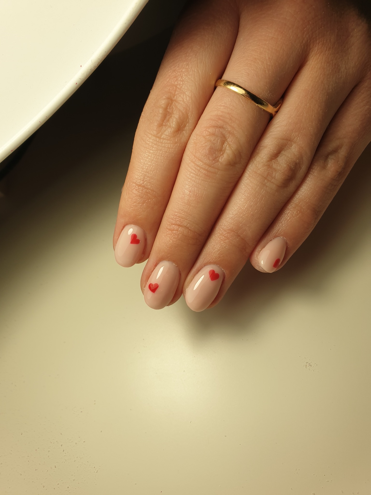 Dłoń z delikatnym, beżowym manicure z małymi czerwonymi serduszkami na każdym paznokciu, ozdobiona złotym pierścionkiem na palcu serdecznym, na jasnym tle.