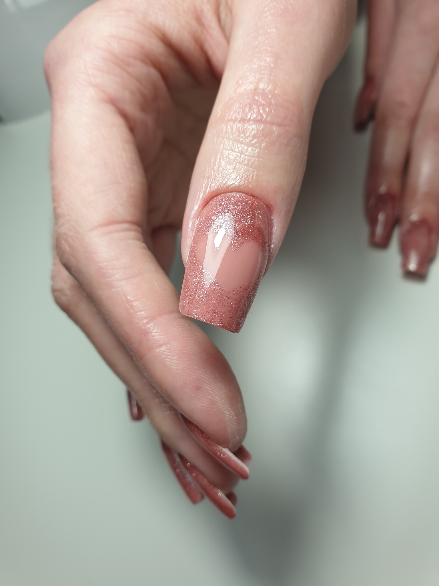 Kwadratowy paznokieć z efektem brokatu i serca w kolorze cielistego różu, widoczne drobne zmarszczki na skórze dłoni.