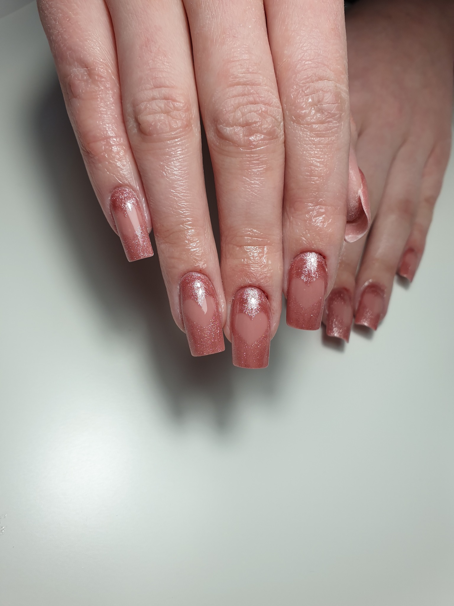 Dłonie z kwadratowymi paznokciami pokrytymi różowym brokatowym lakierem z wzorem serca przy nasadzie na tle białego blatu.