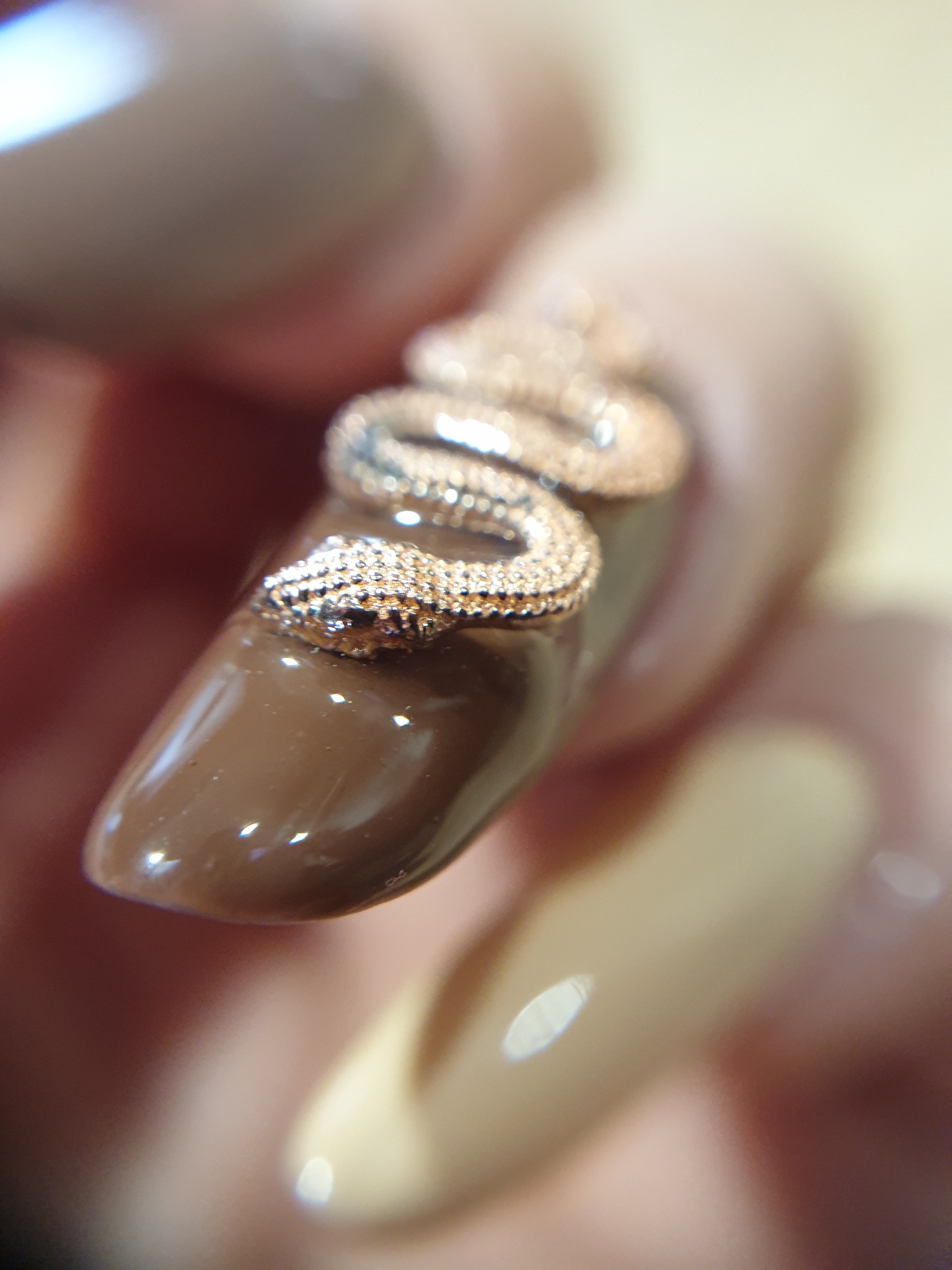 Brązowy, błyszczący paznokieć z wypukłą, złotą ozdobą w kształcie węża, zbliżenie na detal zdobienia.