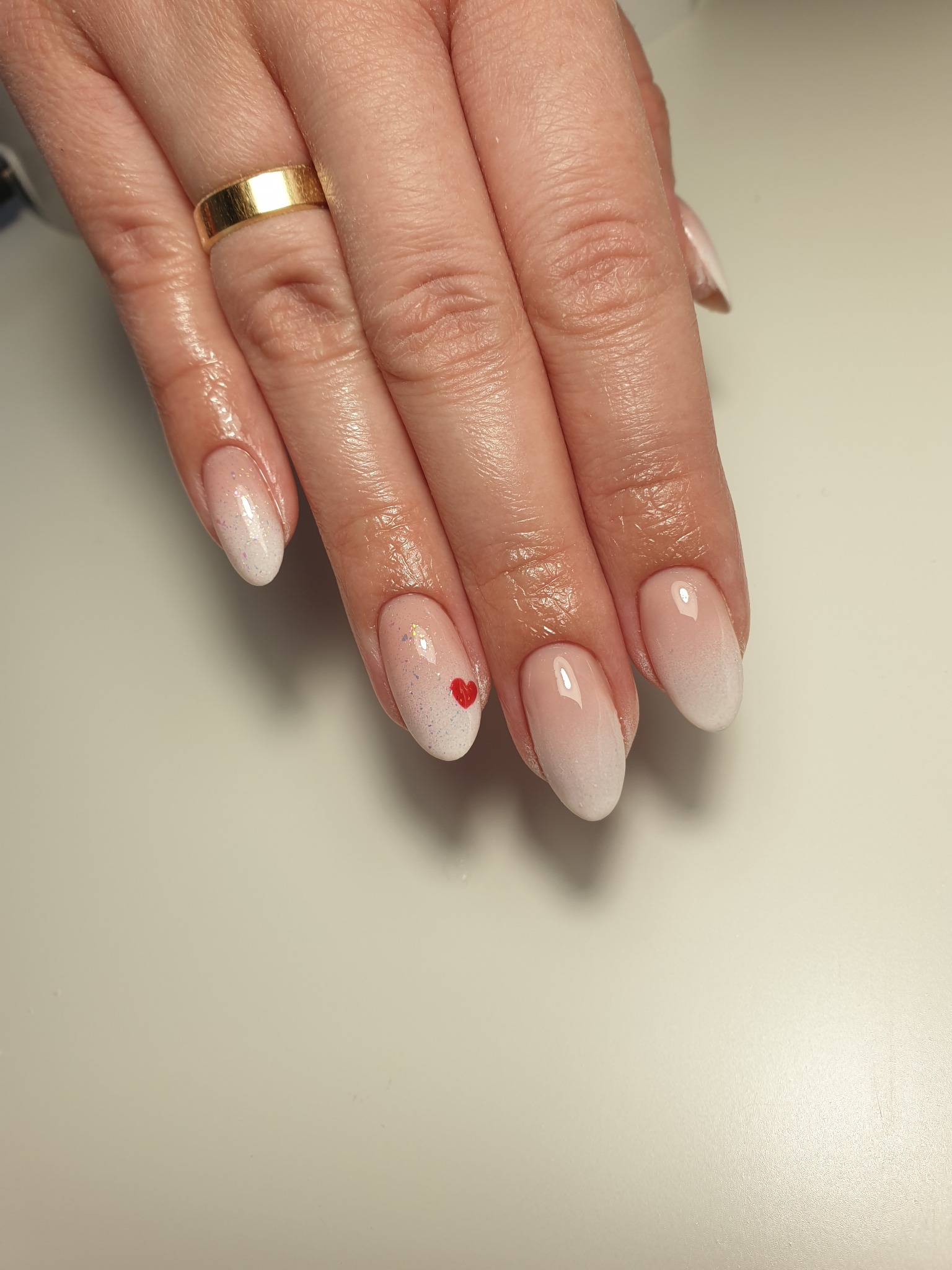 Dłoń z pierścionkiem i subtelnym manicure ombre w odcieniach bieli i różu, jeden paznokieć ozdobiony małym czerwonym serduszkiem.
