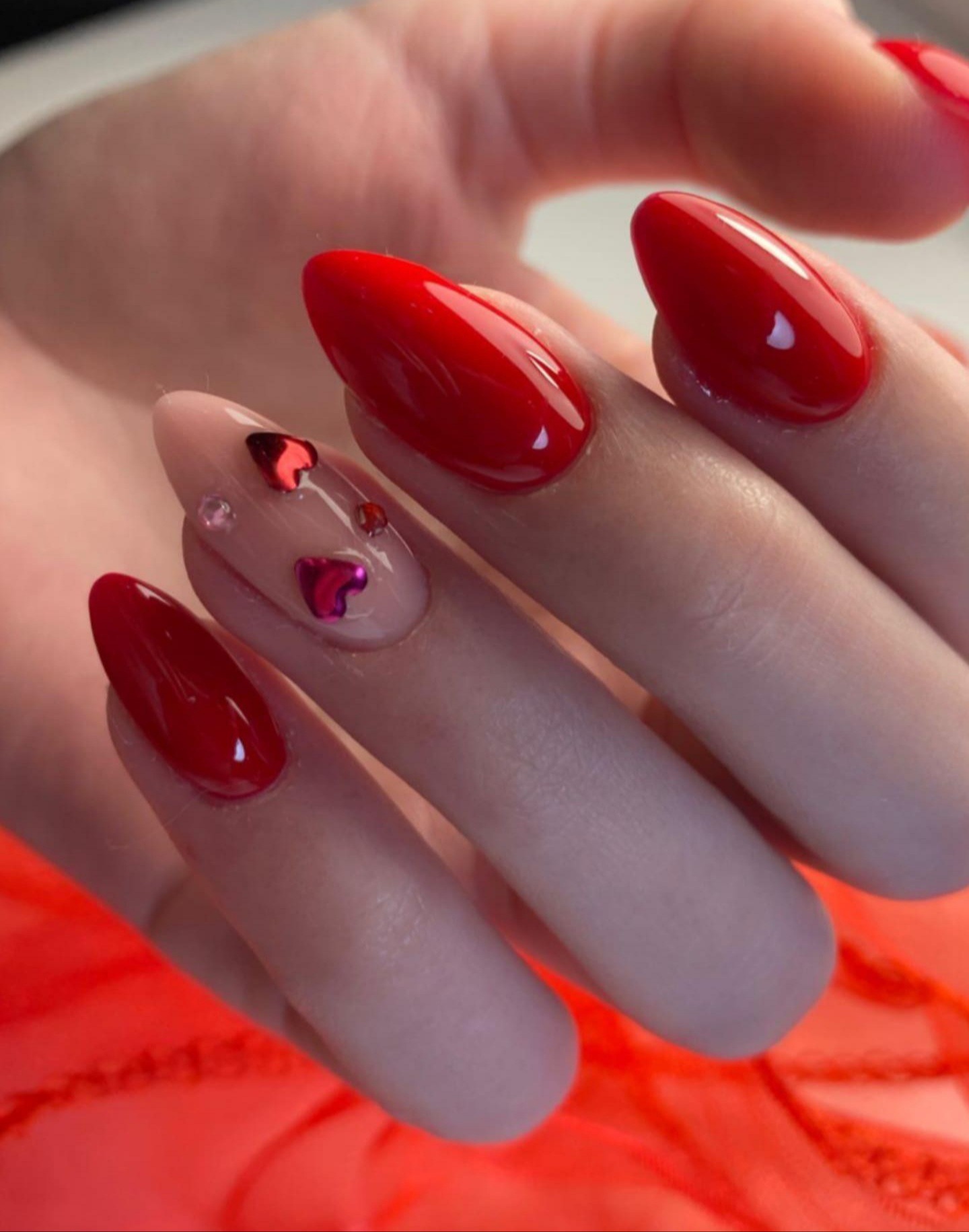 Dłoń z czerwonymi, błyszczącymi paznokciami w kształcie migdała, jeden paznokieć ozdobiony małymi, czerwonymi serduszkami i kryształkami.