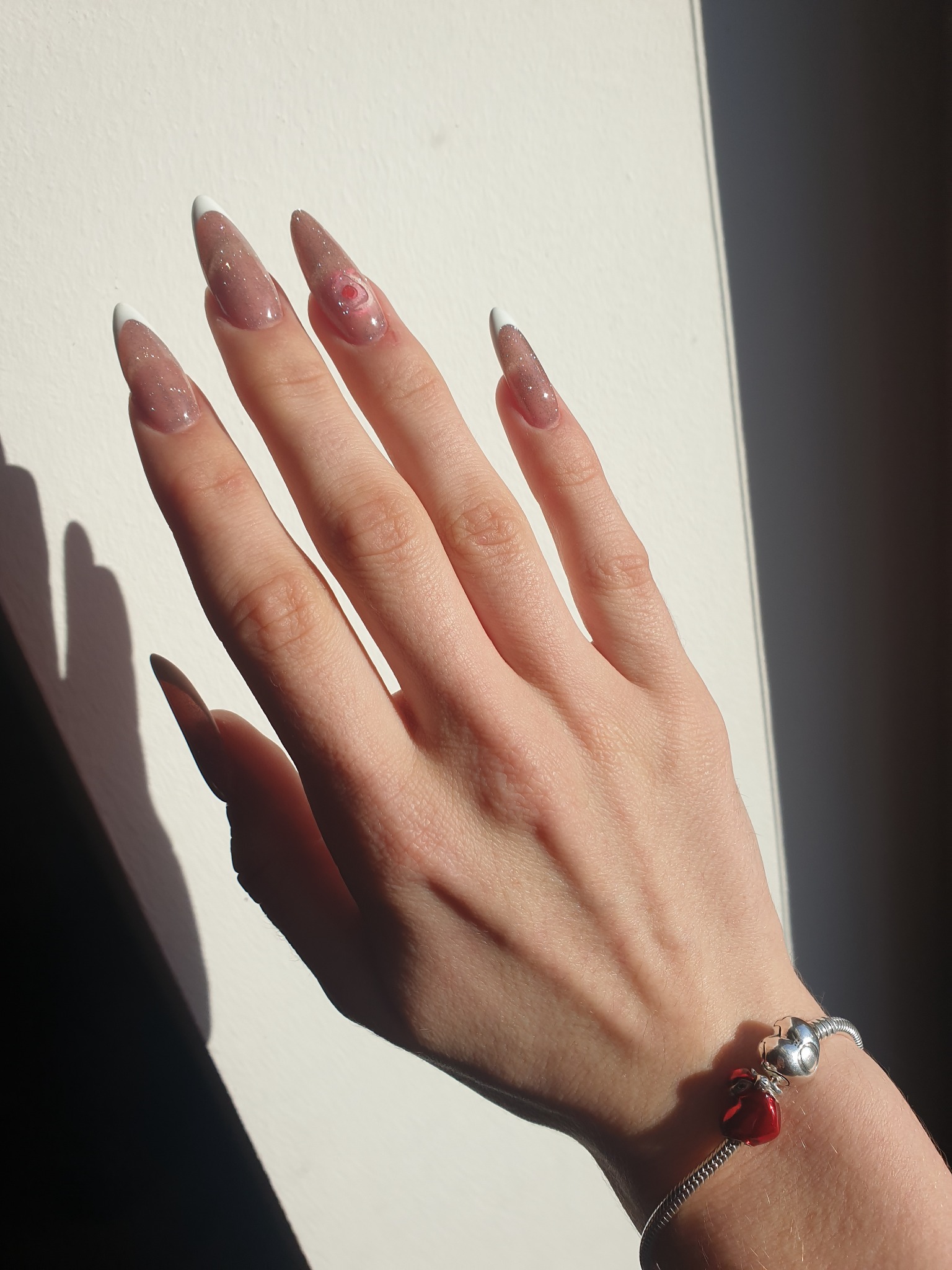 Dłoń z długimi, półprzezroczystymi paznokciami w odcieniach beżu z białymi końcówkami i subtelnym brokatem, jeden paznokieć ozdobiony motywem kwiatowym, na nadgarstku srebrna bransoletka...