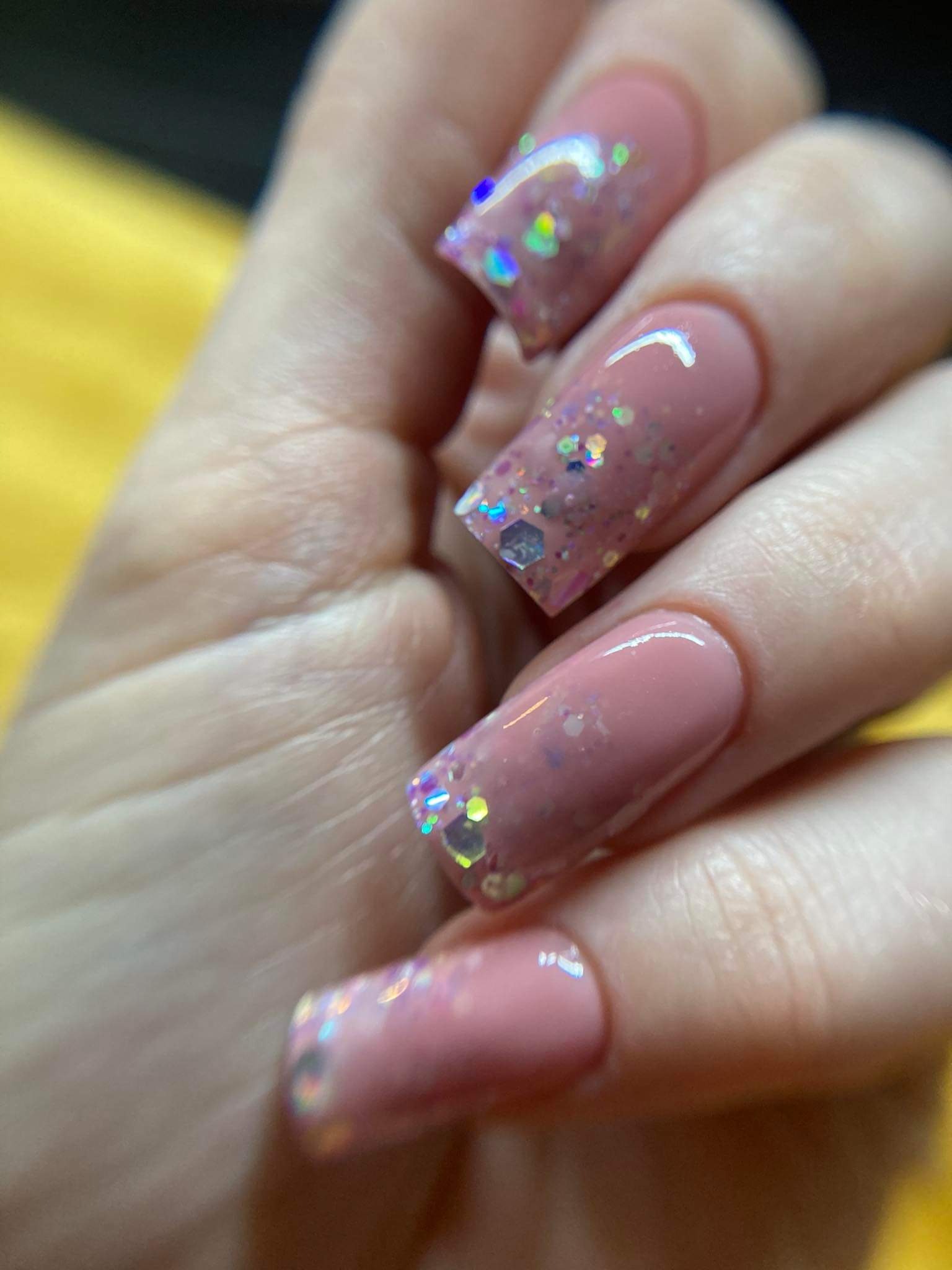 Detaliczne ujęcie dłoni z kwadratowymi paznokciami w odcieniach pudrowego różu, zdobionymi brokatem holograficznym i delikatnymi refleksami światła.