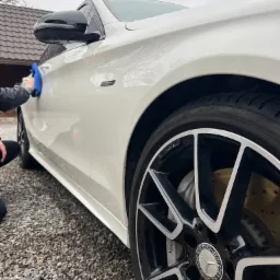 Osoba przeciera niebieską ściereczką biały samochód marki Mercedes-Benz AMG z czarnymi felgami na tle żwirowego podłoża i domu.