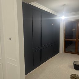 Myjnia Parowa Wash - Wnętrze w trakcie malowania: ciemna ściana z dekoracyjnymi panelami, białe listwy przypodłogowe i ościeżnice, widoczne wiaderka z farbą na podłodze.