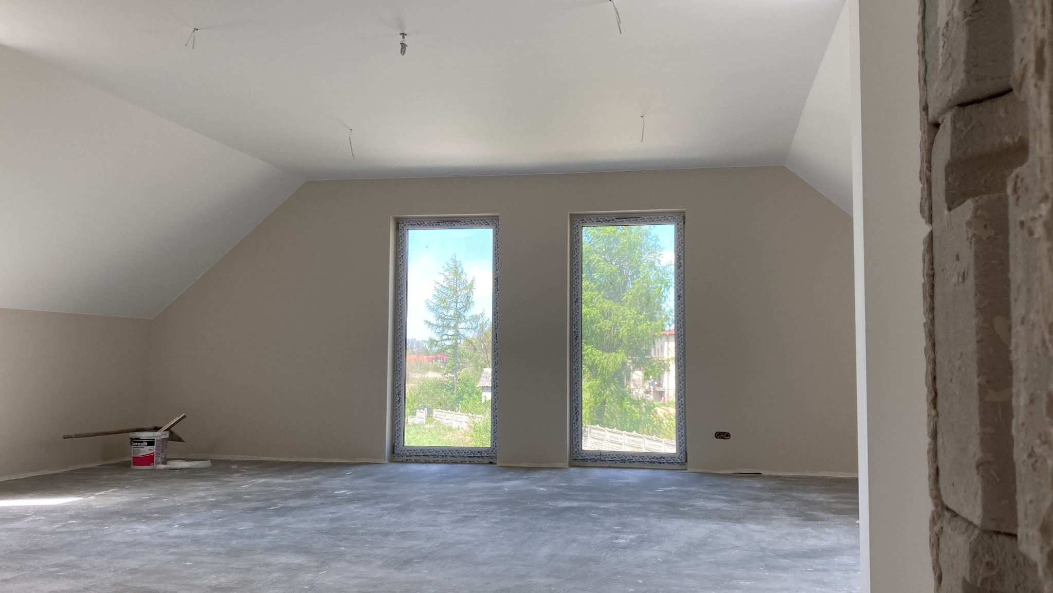 Pokój na poddaszu po szpachlowaniu i gruntowaniu, widoczne dwa okna z widokiem na zieleń, ślady po szpachli na podłodze, wiadro z farbą i narzędzia malarskie przy ścianie.