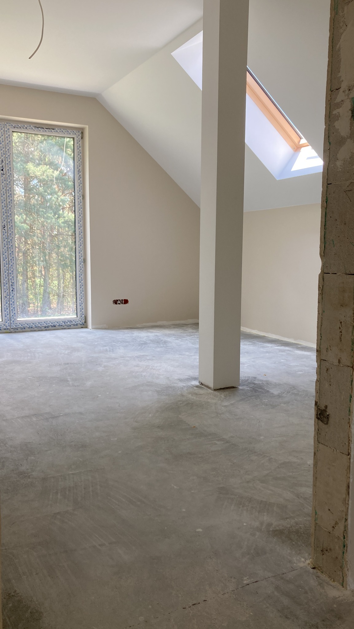 Surowe wnętrze poddasza z oknem dachowym, świeżo pomalowane ściany na jasny beż, widoczne ślady po szpachlowaniu na podłodze, surowa ściana w przejściu.