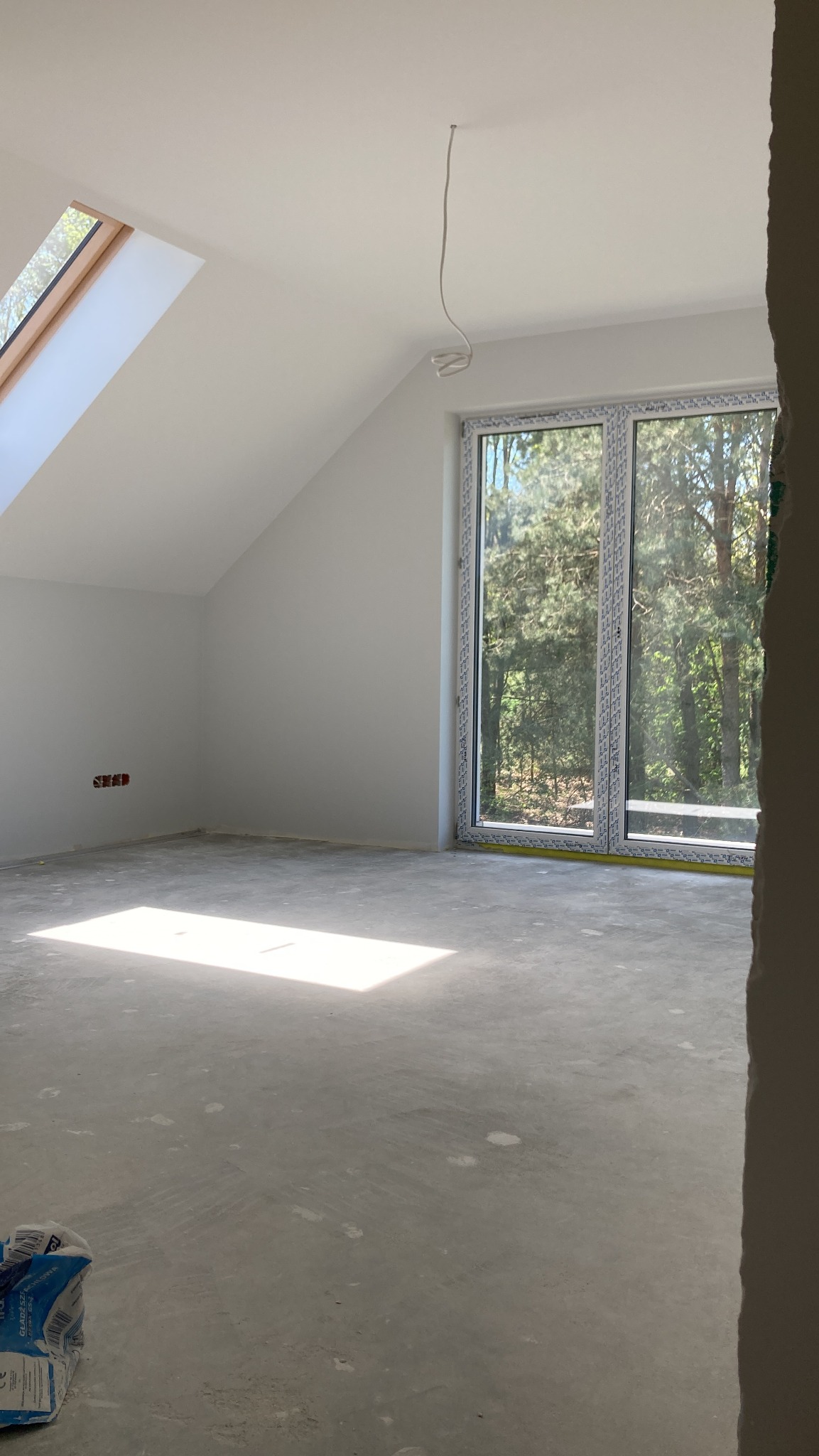 Poddasze w stanie deweloperskim z oknem dachowym, dużym oknem fasadowym z widokiem na drzewa i workiem z zaprawą gipsową w rogu.