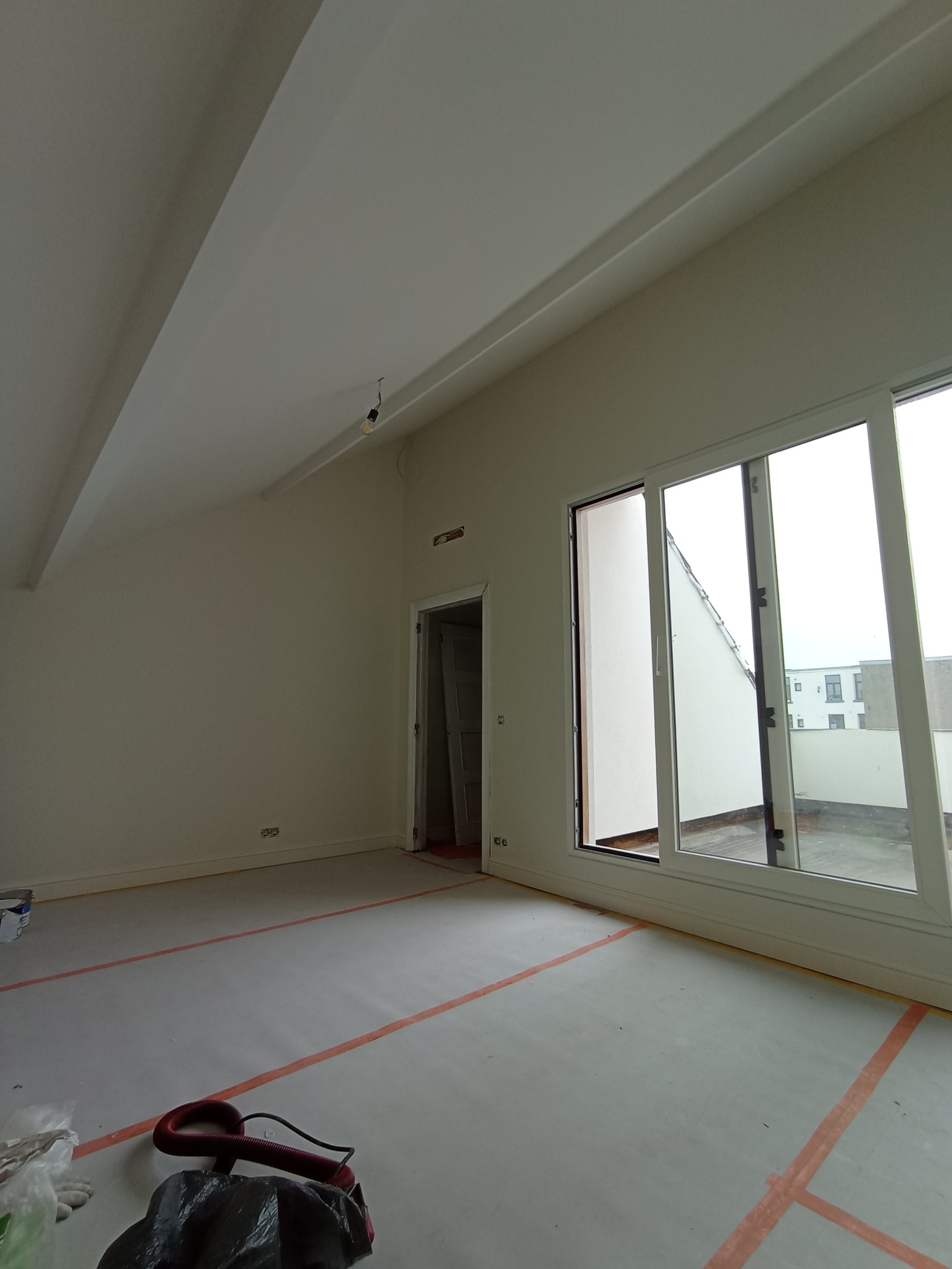 Pokój na poddaszu po szpachlowaniu i przed malowaniem, zabezpieczony pomarańczową taśmą, z widocznymi oknami balkonowymi i sprzętem do sprzątania.