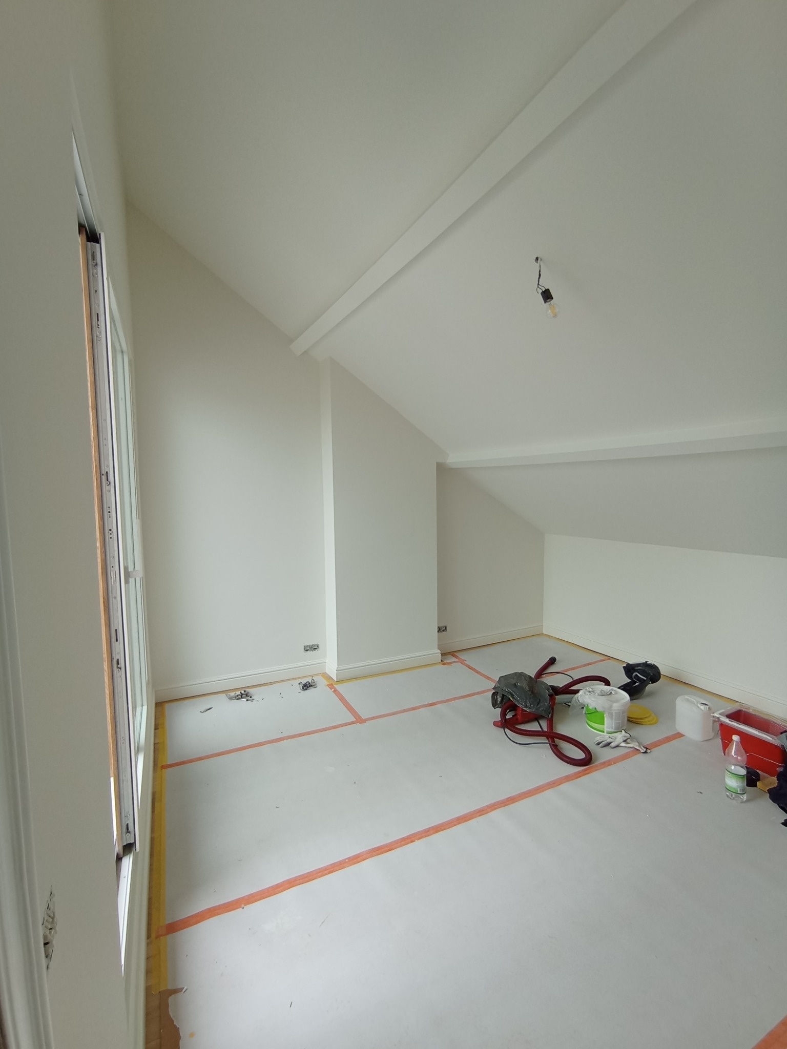 Pokój ze skośnym sufitem po świeżym pomalowaniu na biało, zabezpieczony folią malarską i taśmą. Widoczne narzędzia malarskie i odkurzacz.