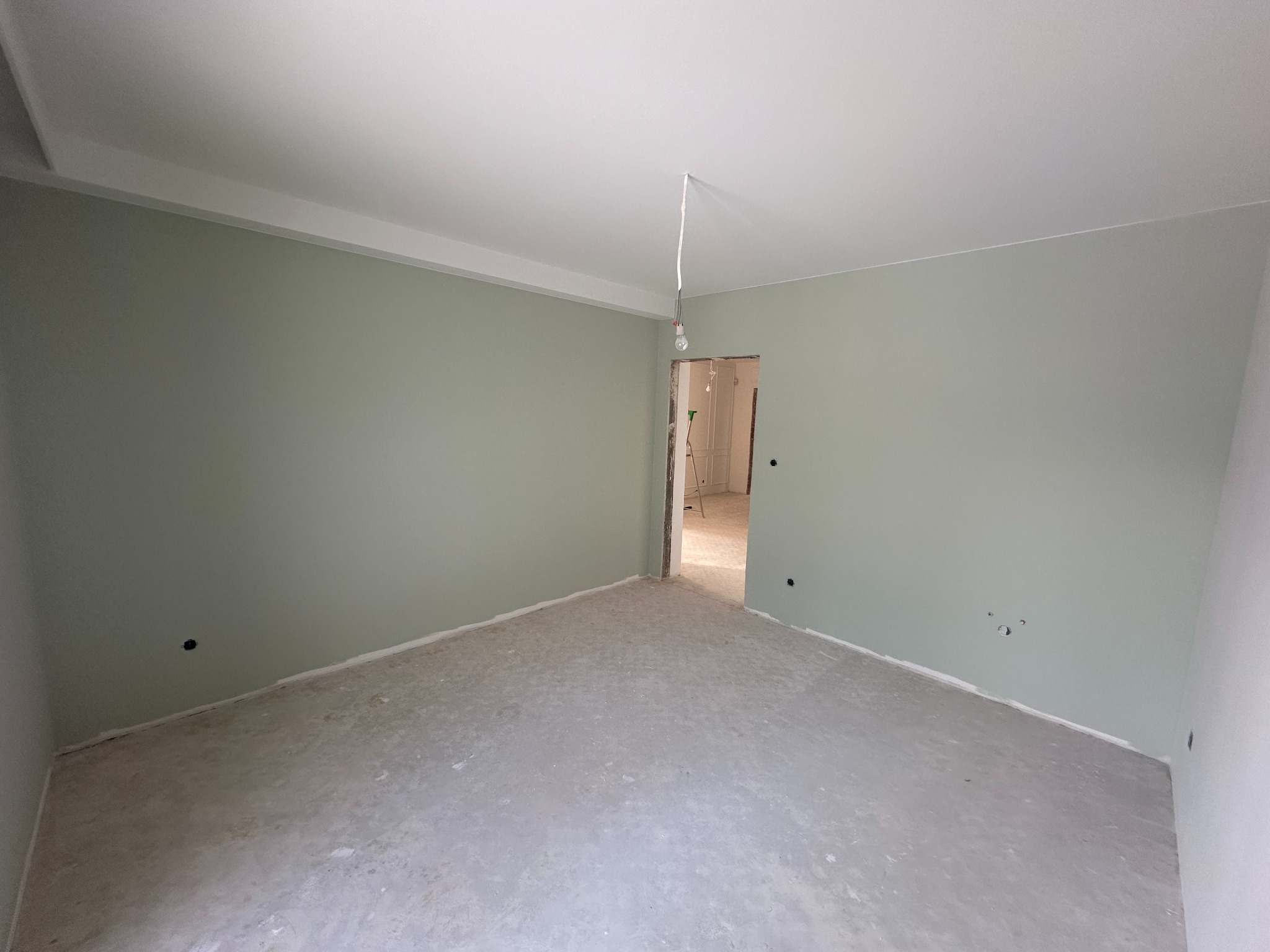 Pusty pokój po malowaniu ścian na delikatny odcień zieleni, widoczne ślady po gładzi na podłodze i wystająca żarówka z sufitu.