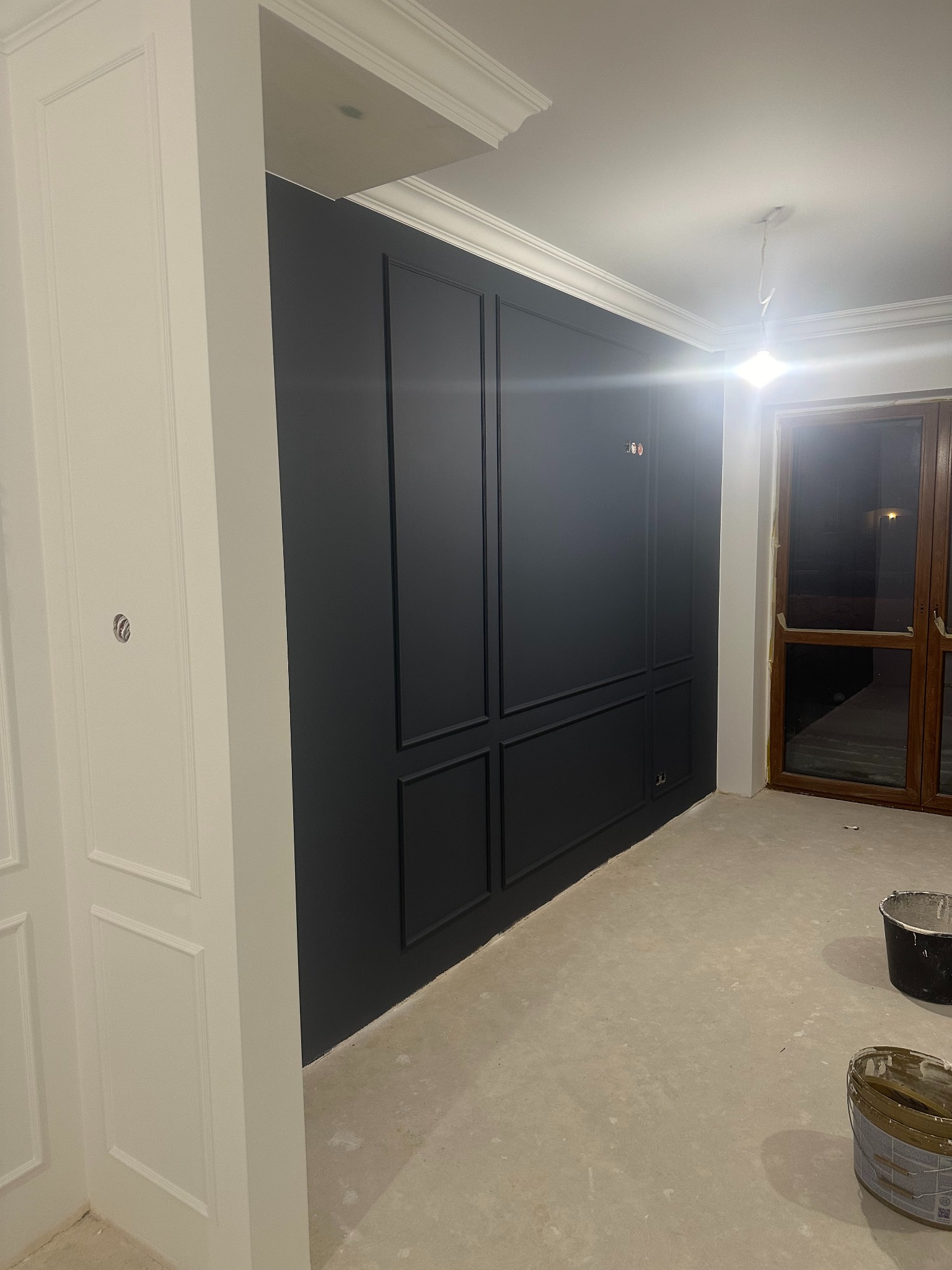 Wnętrze w trakcie malowania: ciemna ściana z dekoracyjnymi panelami, białe listwy przypodłogowe i ościeżnice, widoczne wiaderka z farbą na podłodze.