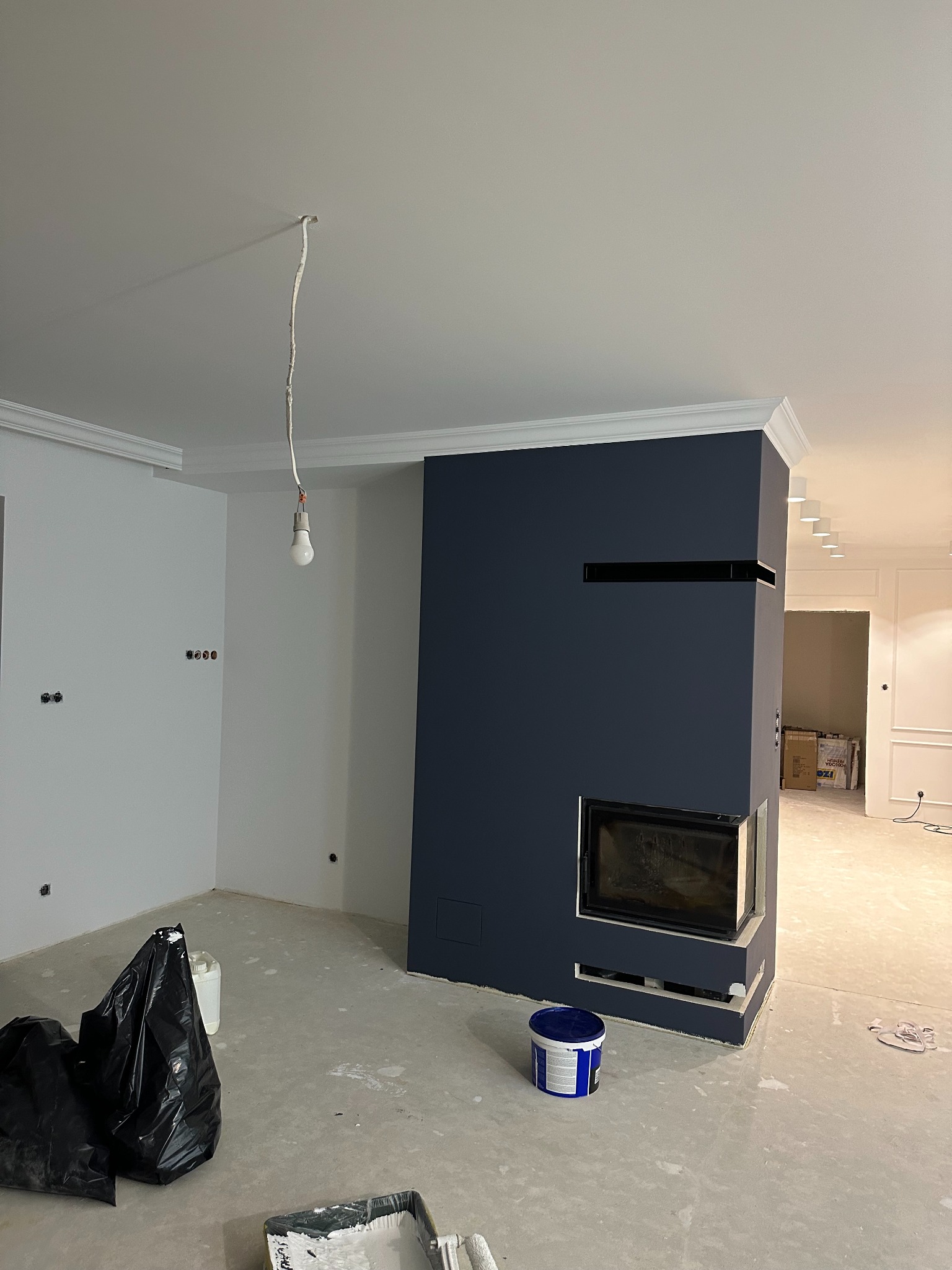 Wnętrze w trakcie remontu: ściany częściowo pomalowane na biało i ciemny granat, widoczny kominek, wisząca żarówka, zakryte folią podłogi i puszka z farbą.