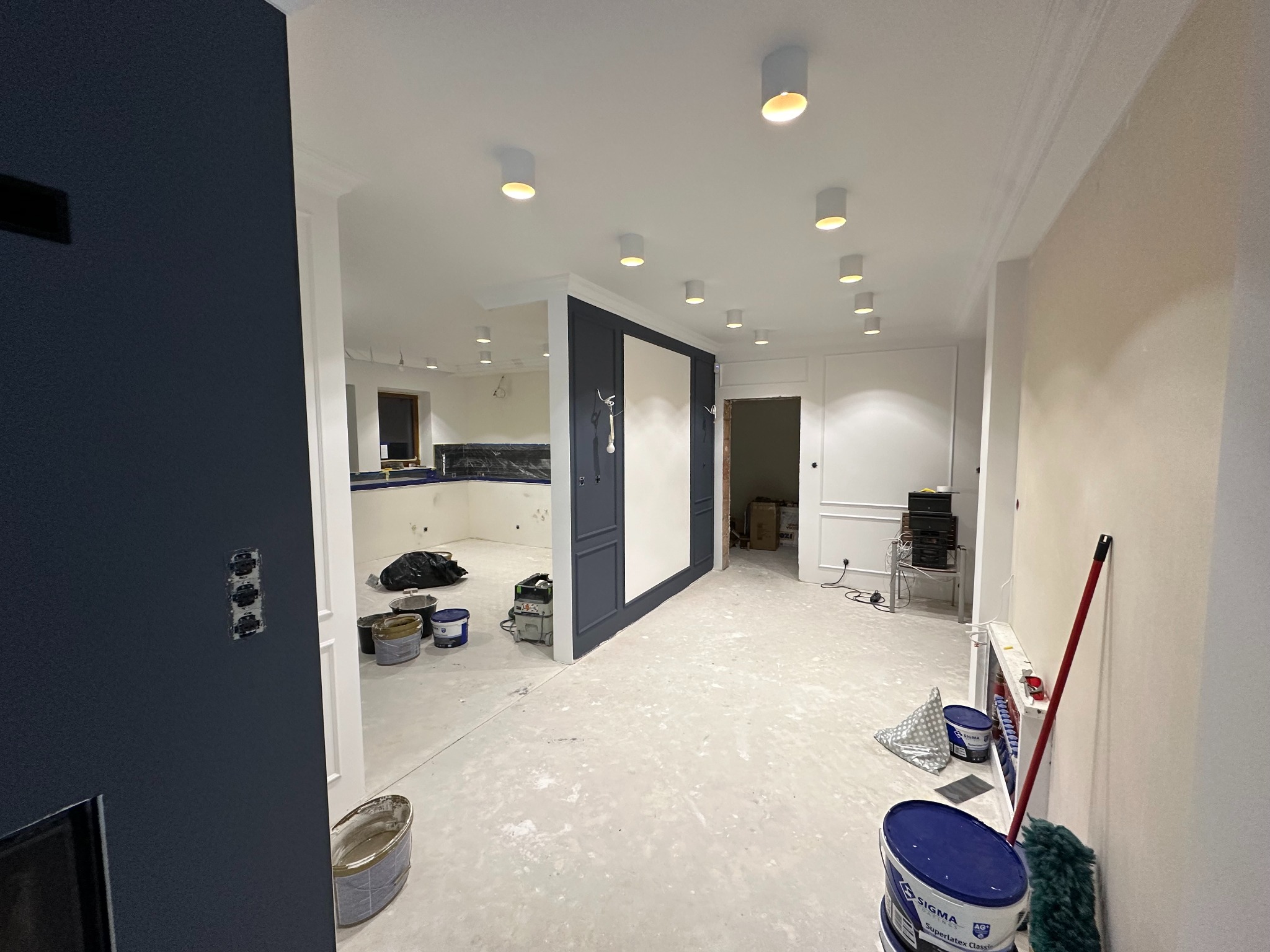 Wnętrze w trakcie prac wykończeniowych: widoczne puszki z farbą, wałek malarski oparty o ścianę, ślady farby na podłodze, ściany częściowo pomalowane na ciemny kolor, a częściowo przygotowane...