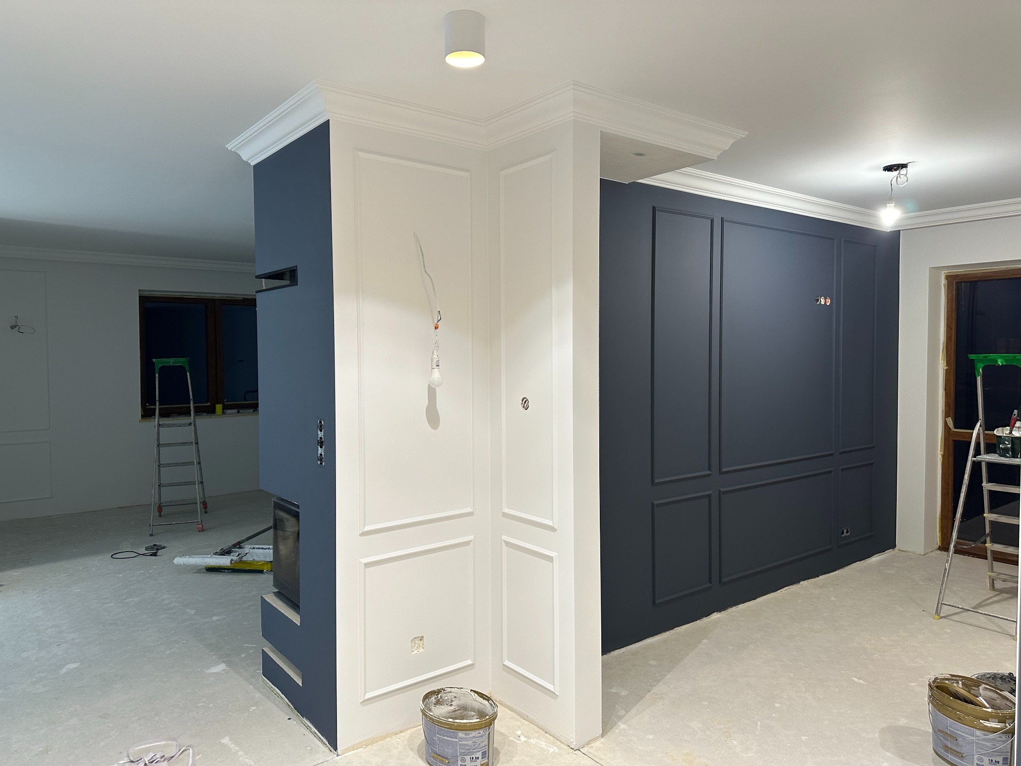 Wnętrze w trakcie prac wykończeniowych: ściany w kolorze granatowym z dekoracyjnymi listwami, białe panele ścienne, widoczne drabiny, puszka z farbą i narzędzia malarskie na podłodze.
