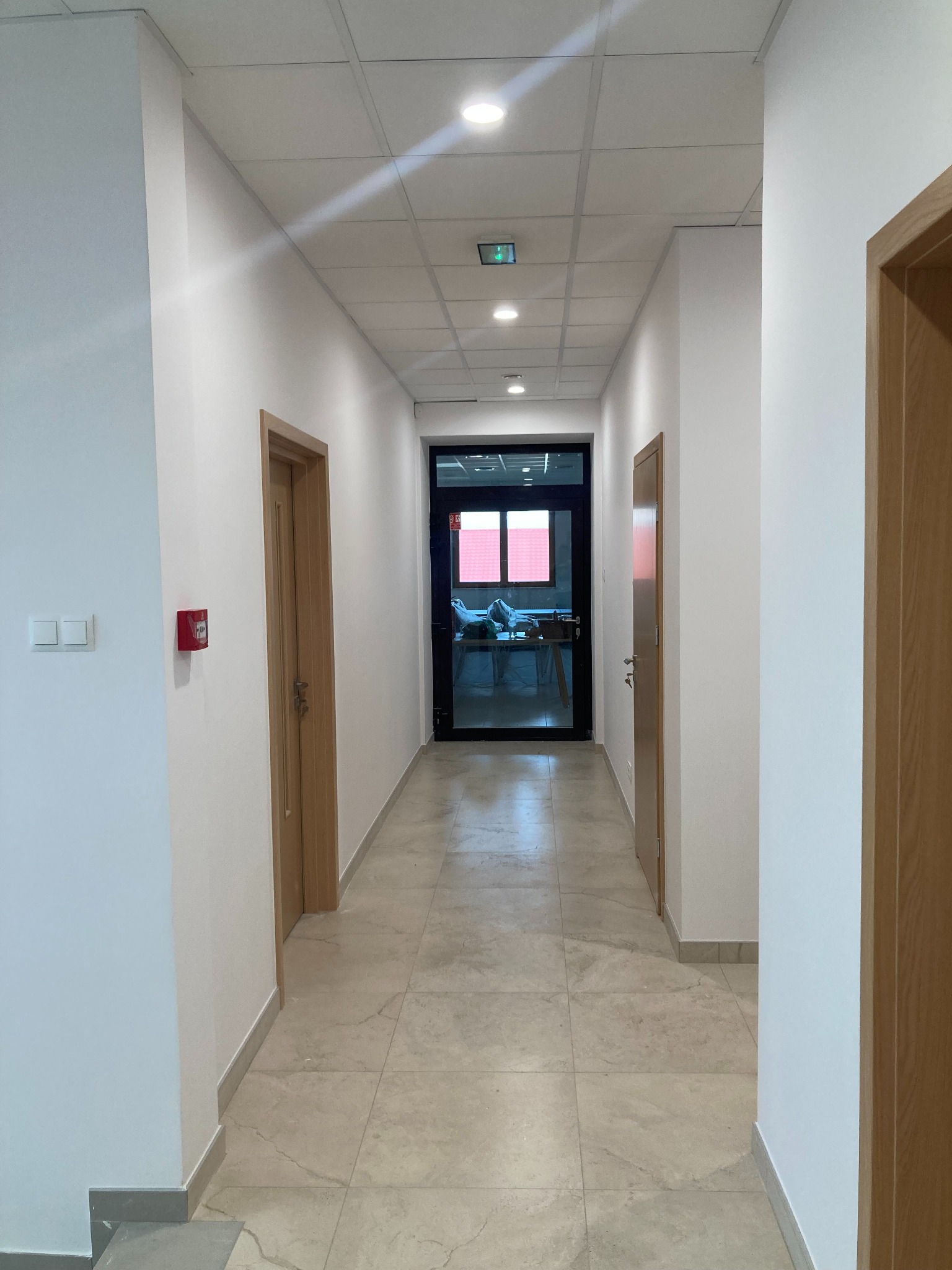 Korytarz biurowy z białymi ścianami, beżową podłogą z płytek i drewnianymi drzwiami. W głębi widoczne przeszklone drzwi do biura.