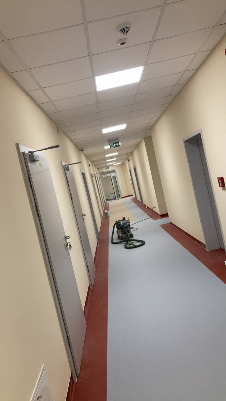 Korytarz biurowy po świeżym pomalowaniu ścian na jasny odcień, z widocznym sprzętem do czyszczenia podłóg i zabezpieczonym fragmentem posadzki na czas prac wykończeniowych.