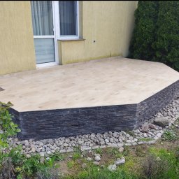 Volodymyr Gonchar - Wykończony taras zewnętrzny z jasnymi płytkami i ciemną okładziną imitującą kamień, otoczony kamieniami i zielenią, z widocznym fragmentem budynku i iglakiem.