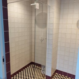 Volodymyr Gonchar - Kabina prysznicowa z białymi płytkami i czarno-białą szachownicą na podłodze, wykończona bordową listwą, widok z góry na deszczownicę.