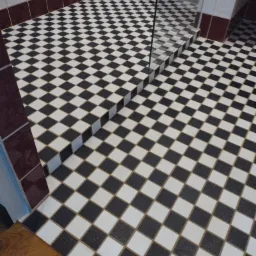 Ukończona podłoga łazienki wyłożona czarno-białą szachownicą, z widocznym fragmentem szklanej kabiny prysznicowej i brązowym pasem płytek przy ścianie.