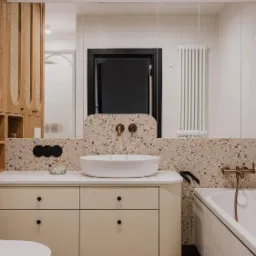 Nowoczesna łazienka z umywalką nablatową, beżową szafką z czarnymi gałkami, ścianą i podłogą wykończoną terazzo, czarną stolarką drzwiową i złotą armaturą