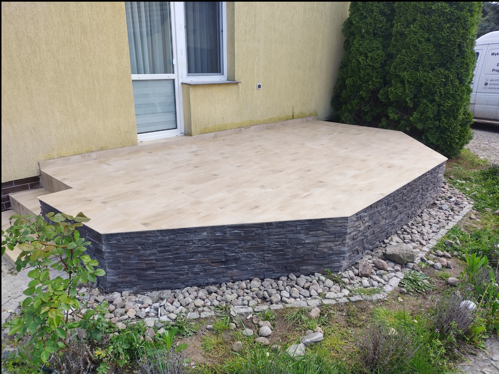 Wykończony taras zewnętrzny z jasnymi płytkami i ciemną okładziną imitującą kamień, otoczony kamieniami i zielenią, z widocznym fragmentem budynku i iglakiem.