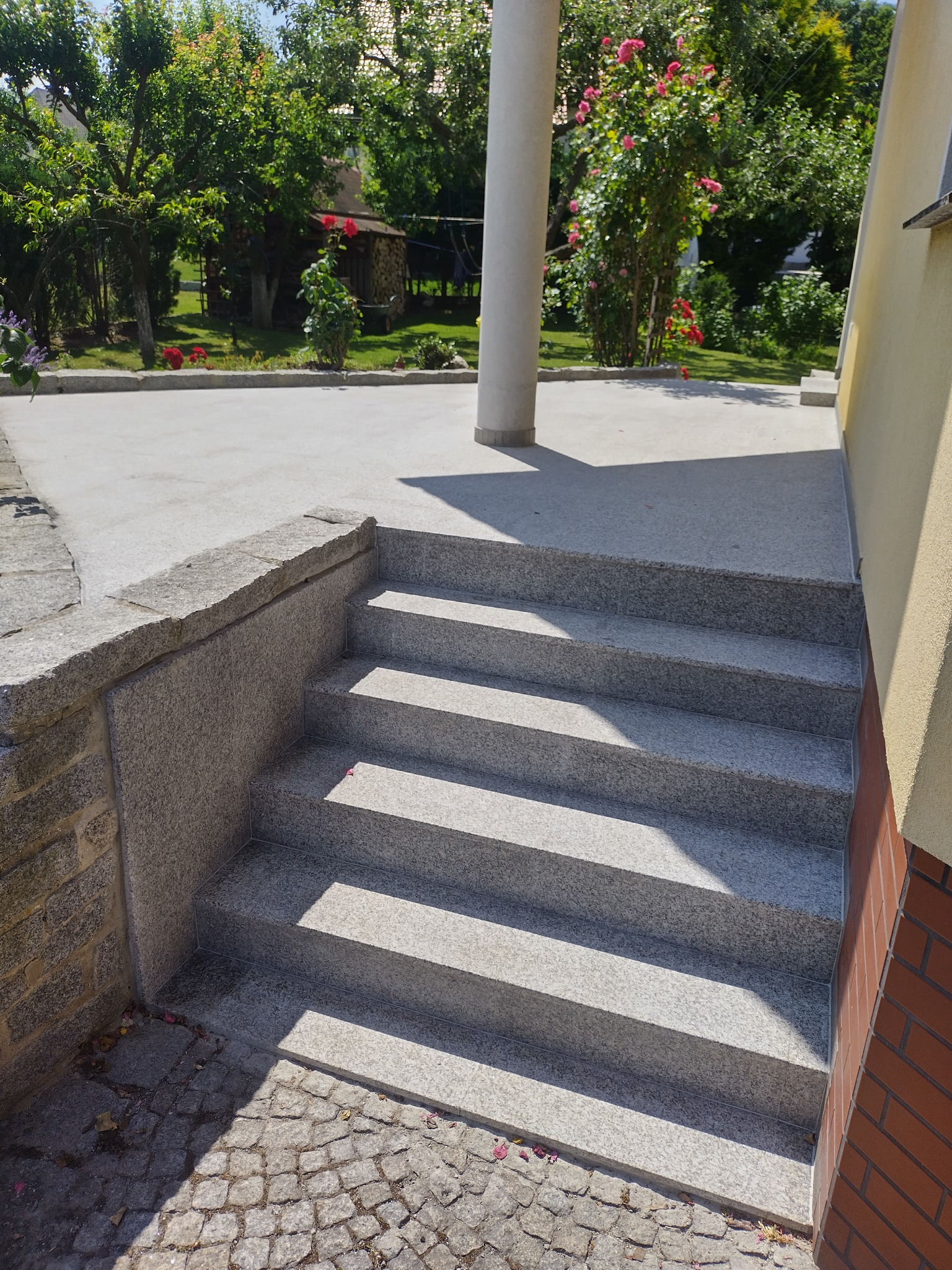 Granitowe schody zewnętrzne prowadzące do domu z kolumną, widok na ogród z różami w tle.