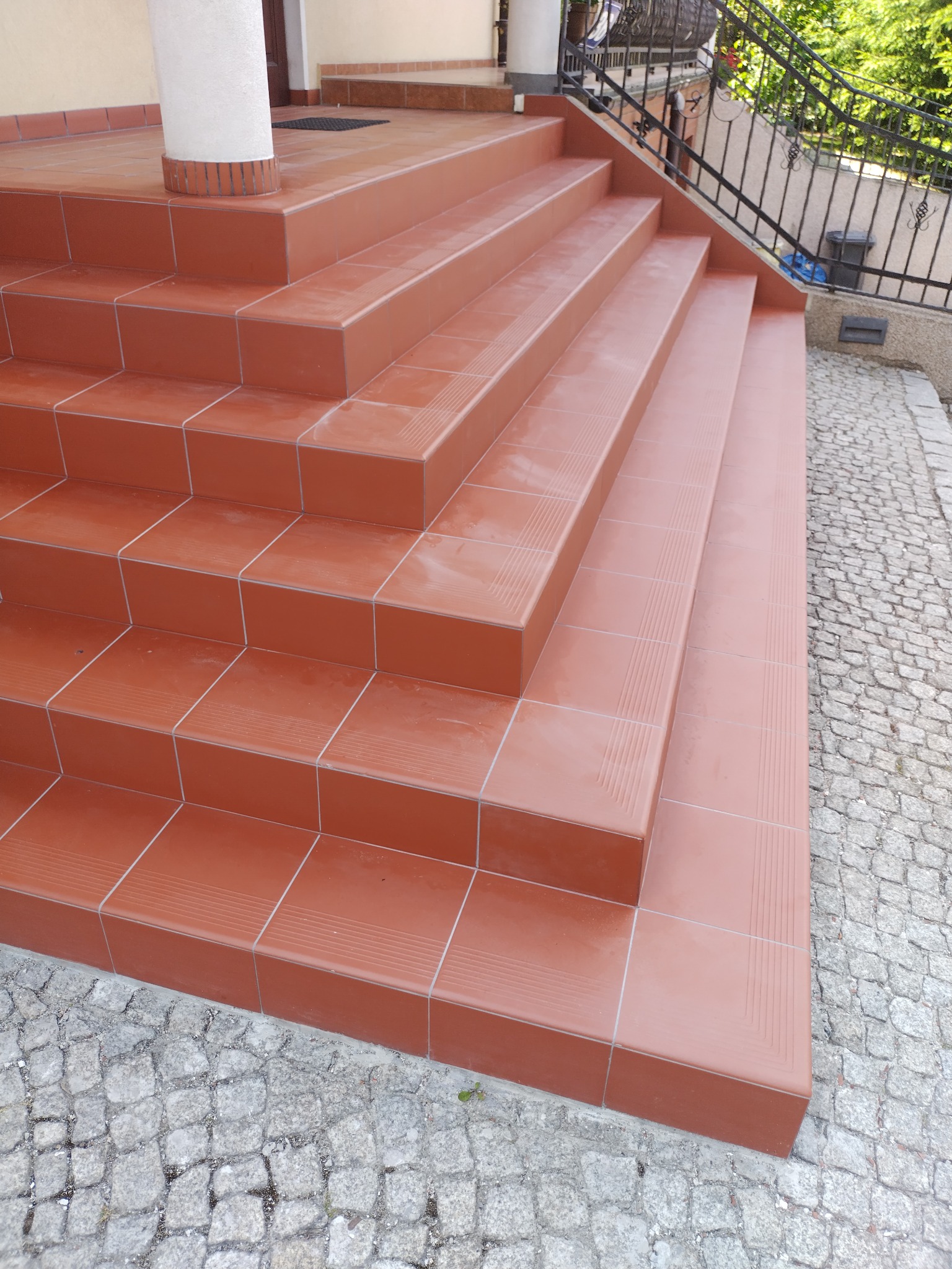 Zewnętrzne schody wejściowe wyłożone terakotą z delikatnym wzorem, obok kostka brukowa i czarna metalowa balustrada.