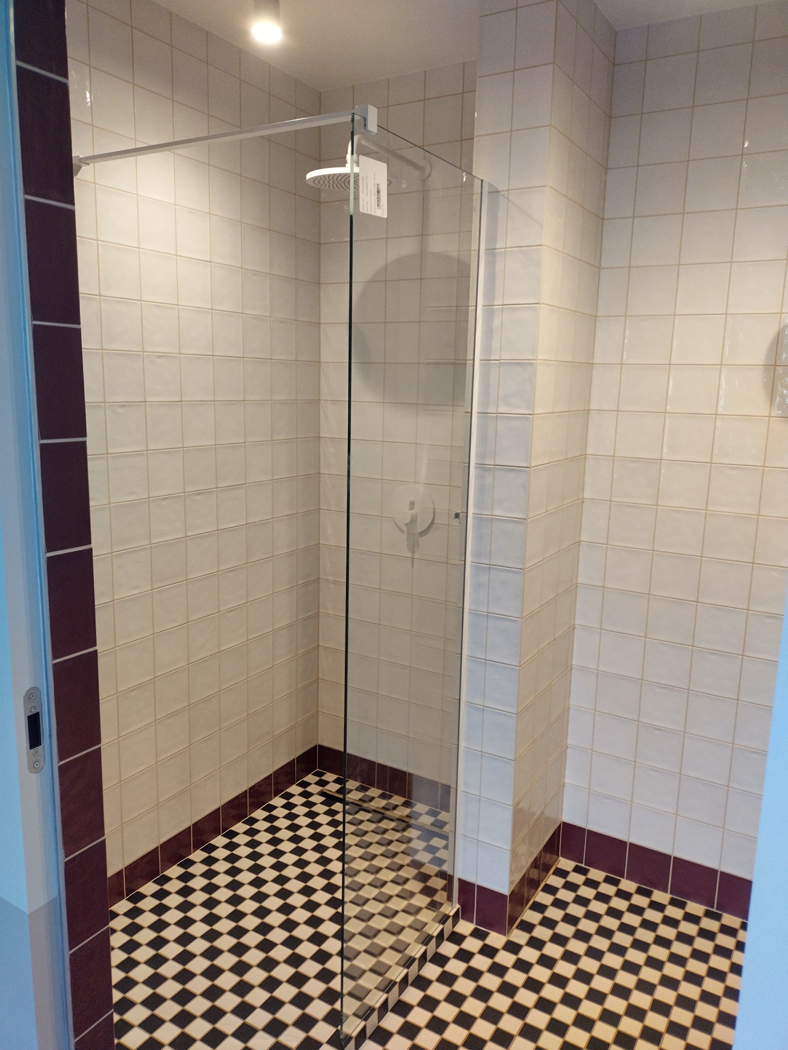 Kabina prysznicowa z białymi płytkami i czarno-białą szachownicą na podłodze, wykończona bordową listwą, widok z góry na deszczownicę.