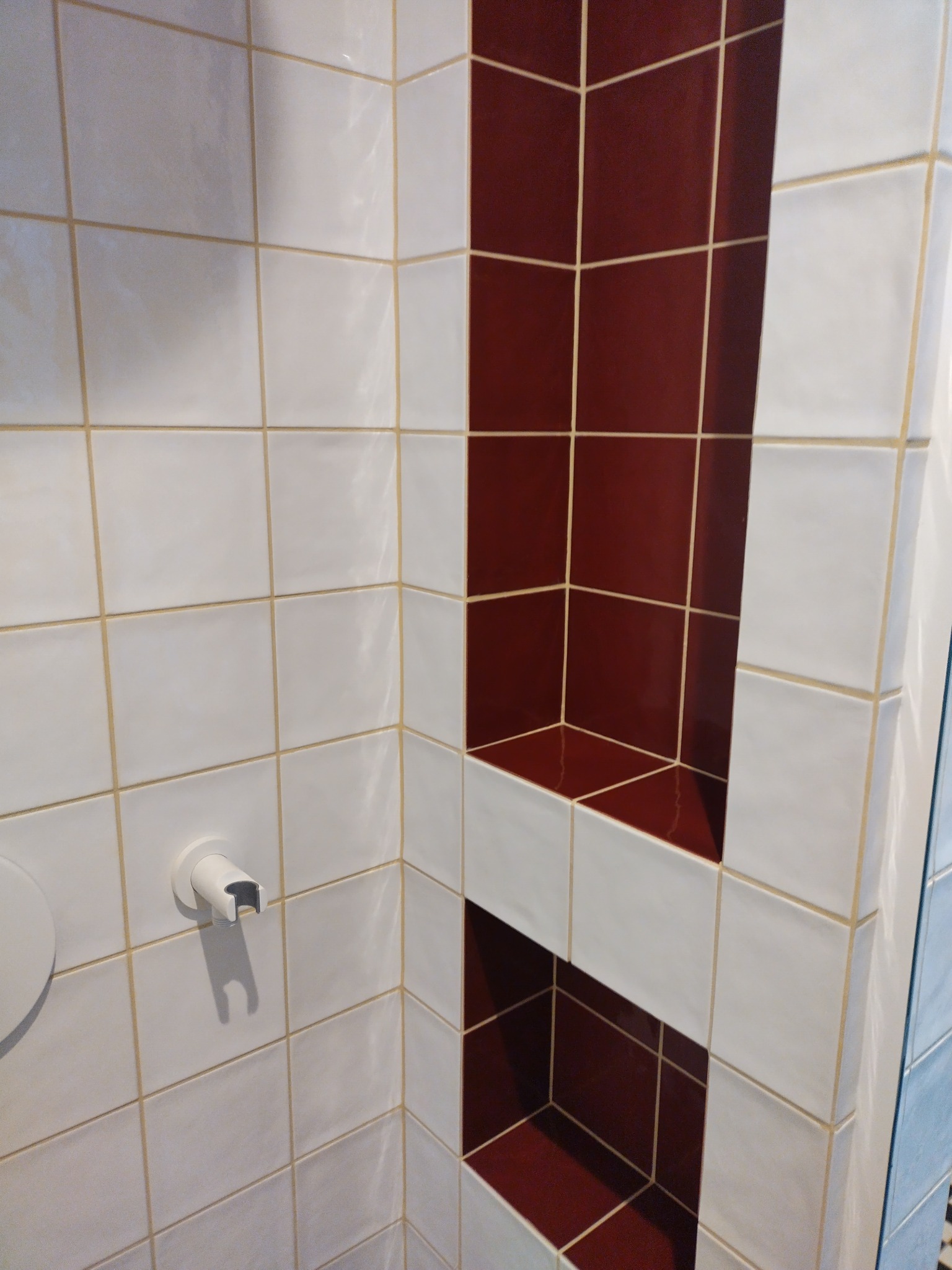 Wnęka w kabinie prysznicowej wyłożona bordowymi płytkami, otoczona białymi płytkami z jasnobeżową fugą i uchwytem na słuchawkę prysznicową.