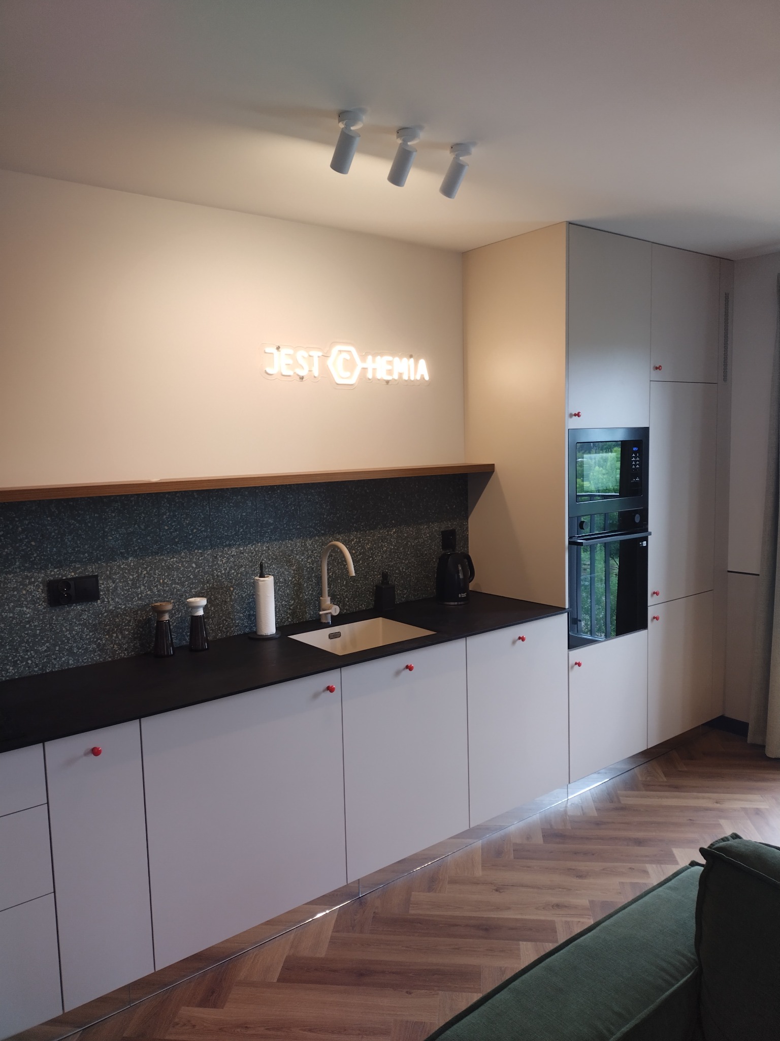 Nowoczesna kuchnia z jasnymi szafkami z czerwonymi uchwytami, czarnym blatem, mozaiką na ścianie i neonowym napisem 'JEST CHEMIA' nad zlewem, oświetlona reflektorami.