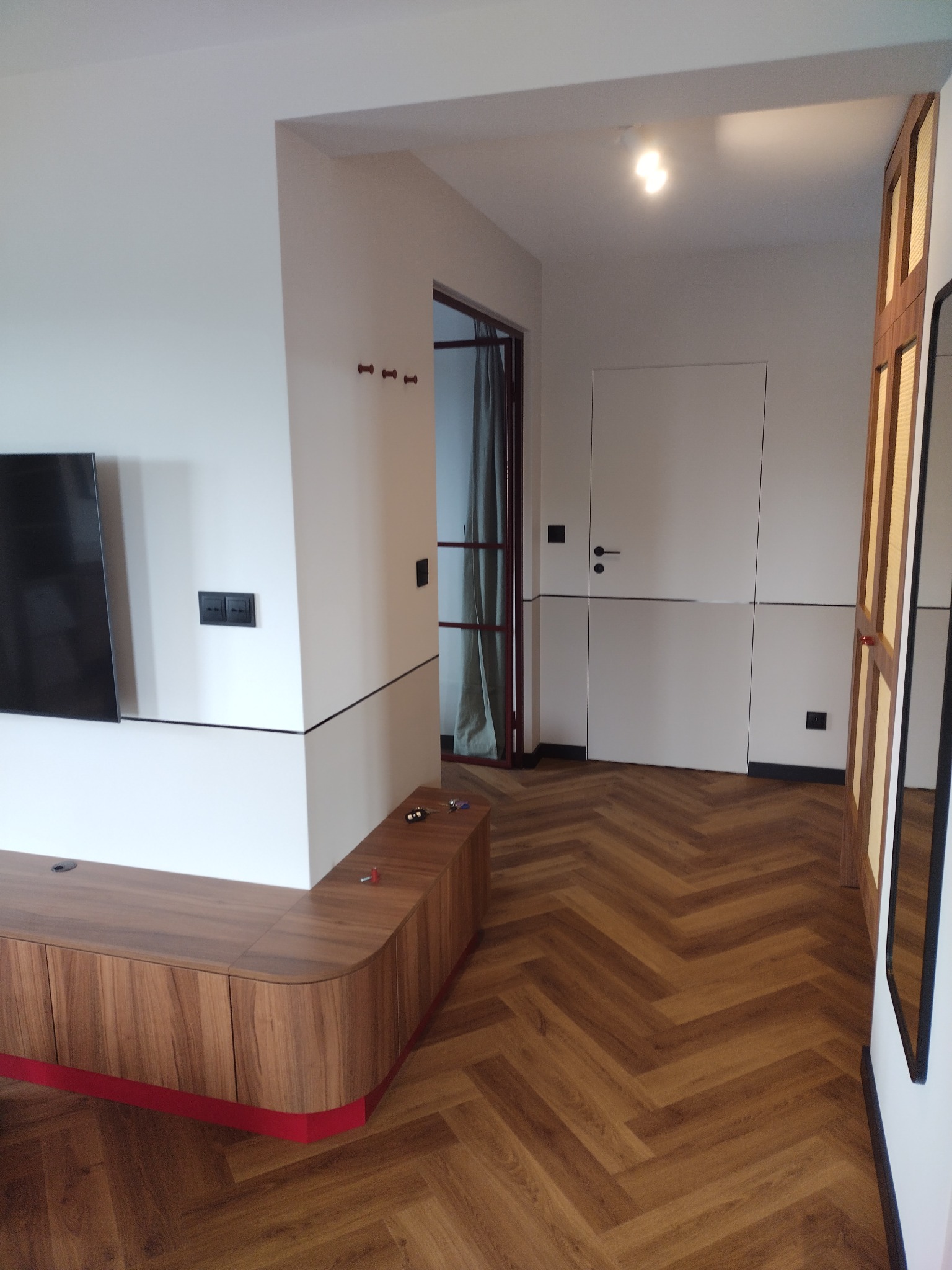 Nowoczesne wnętrze mieszkania z parkietem ułożonym w jodełkę, białe ściany z czarnymi akcentami, drewniana szafka RTV z czerwonym cokołem oraz widoczne drzwi z czerwonymi ramami.