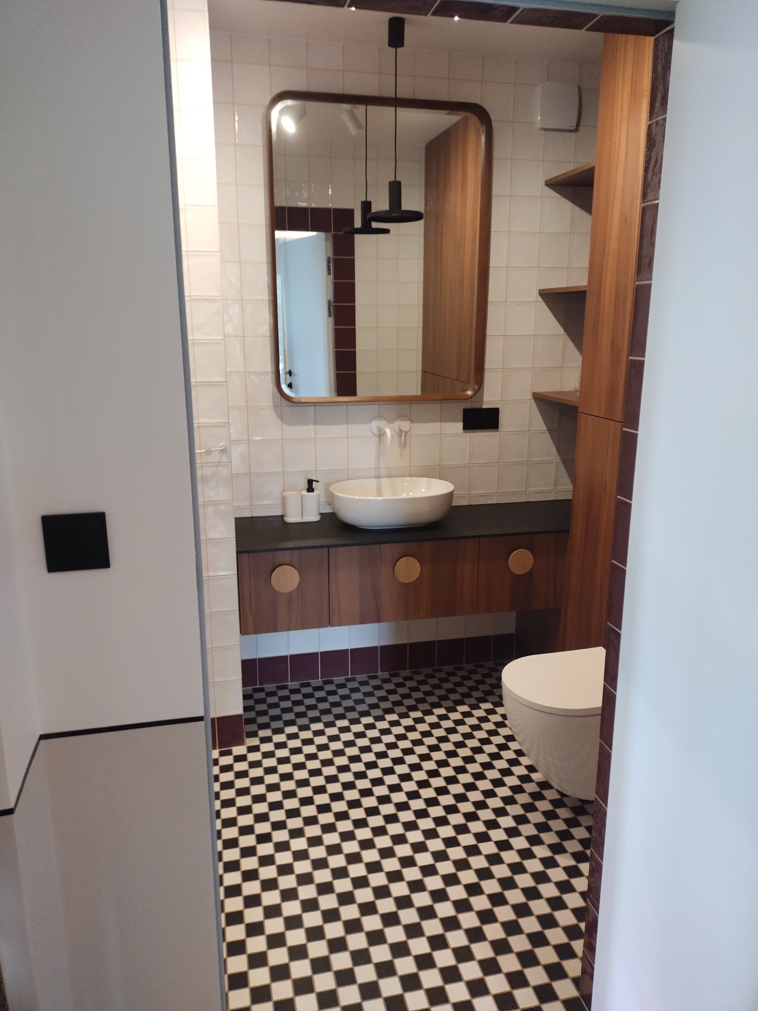 Nowoczesna łazienka z białą umywalką nablatową, lustrem w drewnianej ramie, czarno-białą szachownicą na podłodze i zabudową z drewnianymi szafkami oraz półkami.
